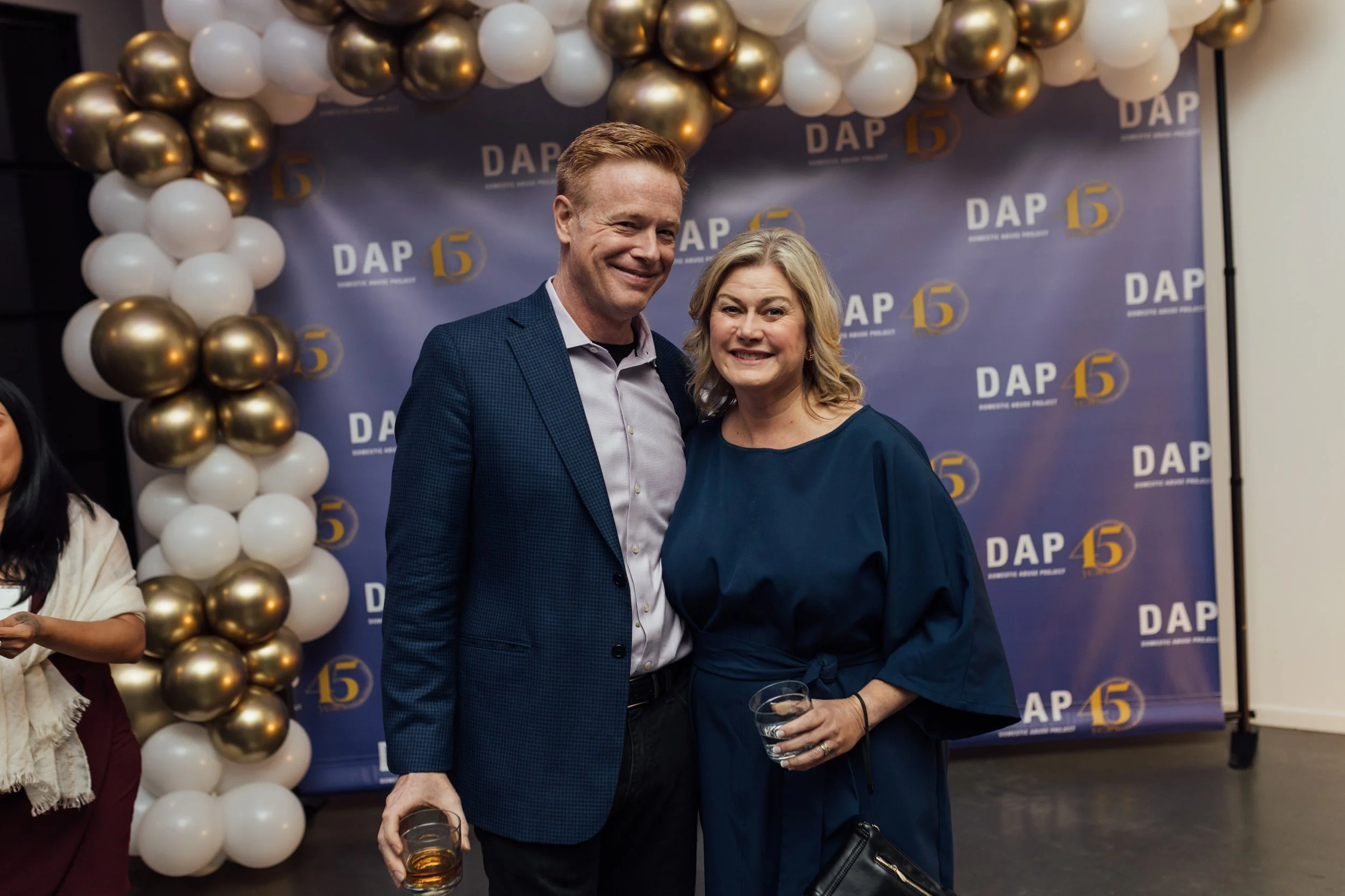 A man and a woman smiling and posing together at a celebration event, holding drinks. The backdrop behind them features the number 15 and the text 'DAP' repeated, with white and gold balloon decorations.