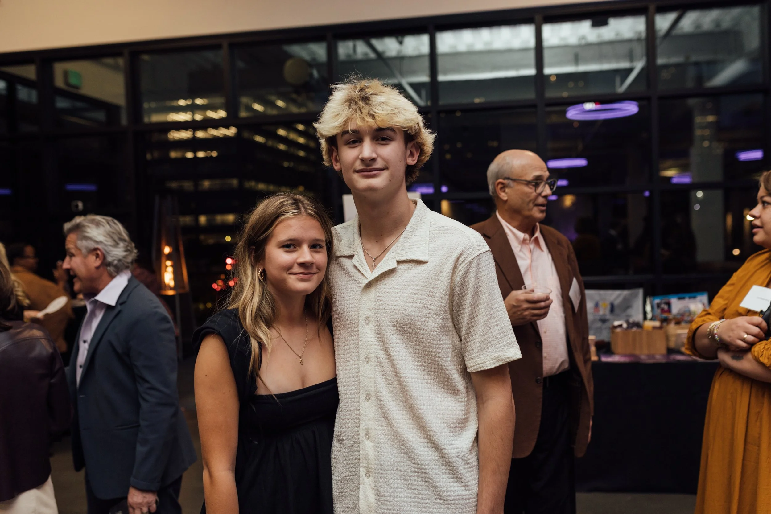 A young man with curly blond hair and a young woman with long light brown hair standing close together at a social event with other attendees in the background.