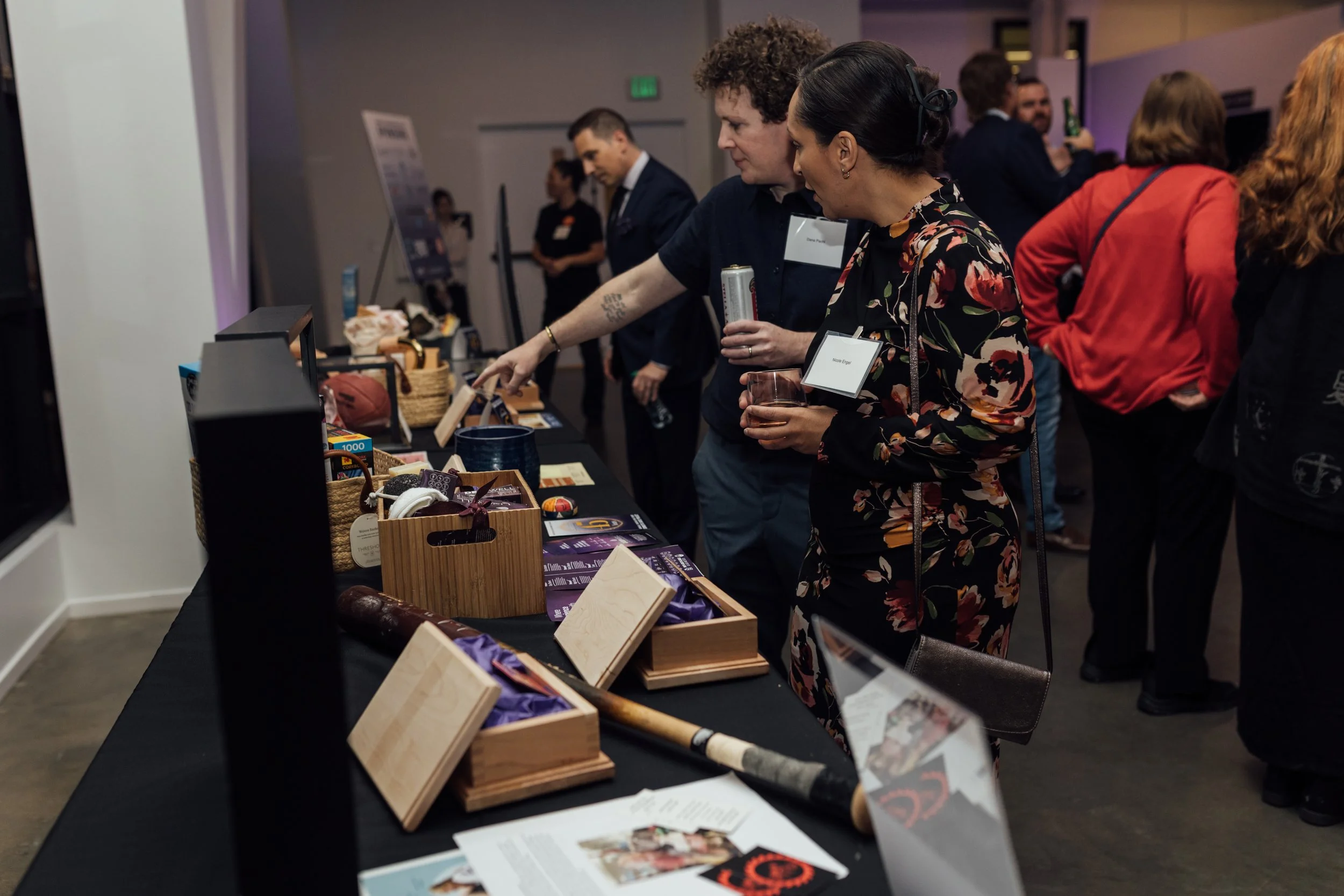 People browsing items displayed on a table at an indoor event or exhibition, with some holding drinks and engaging with the displays.