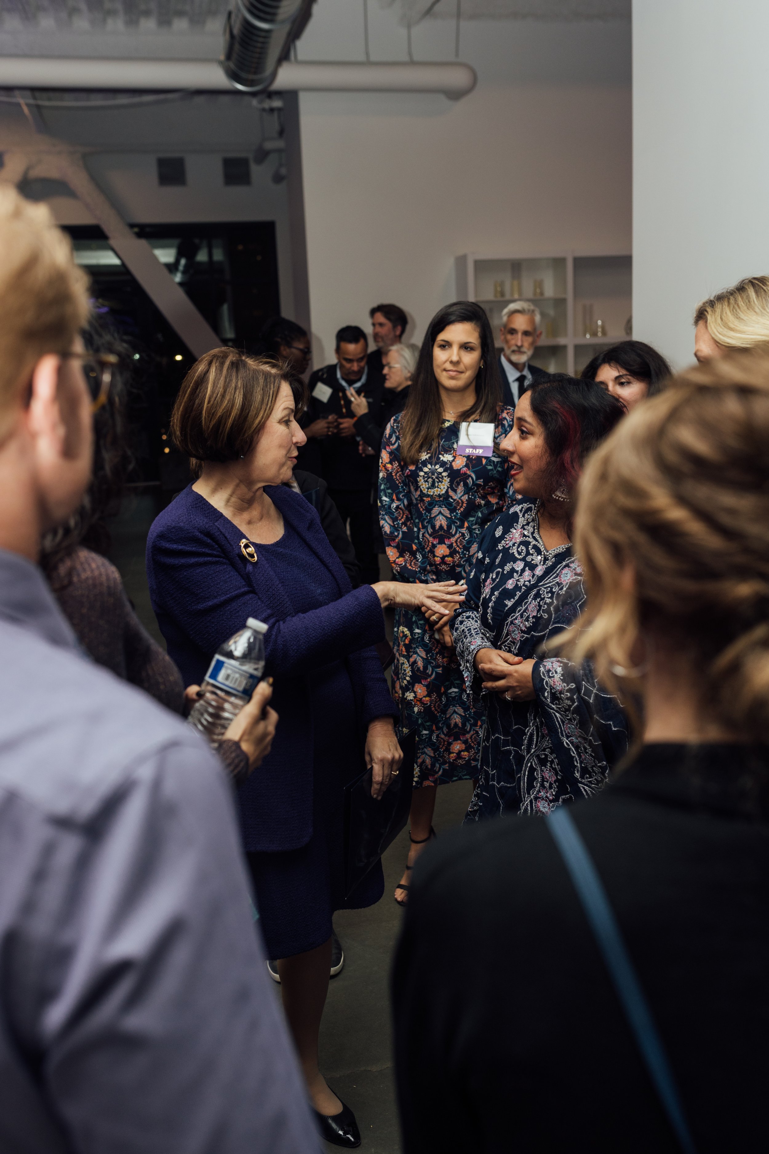 A woman in a dark blue suit is speaking with a woman in a patterned dress at a social gathering, with other people in the background.