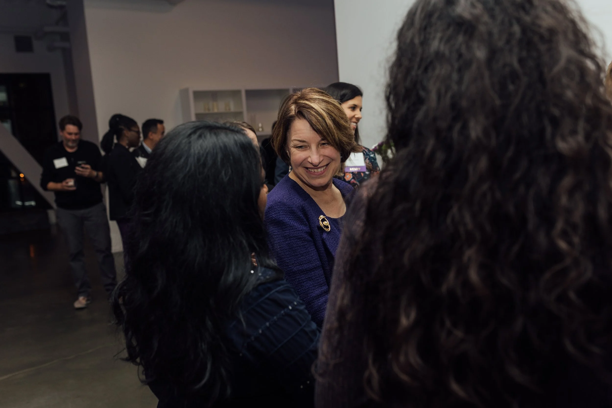 A group of people at a social event, with a woman in a purple blazer smiling and talking to others in the foreground, and others mingling in the background inside a modern room.