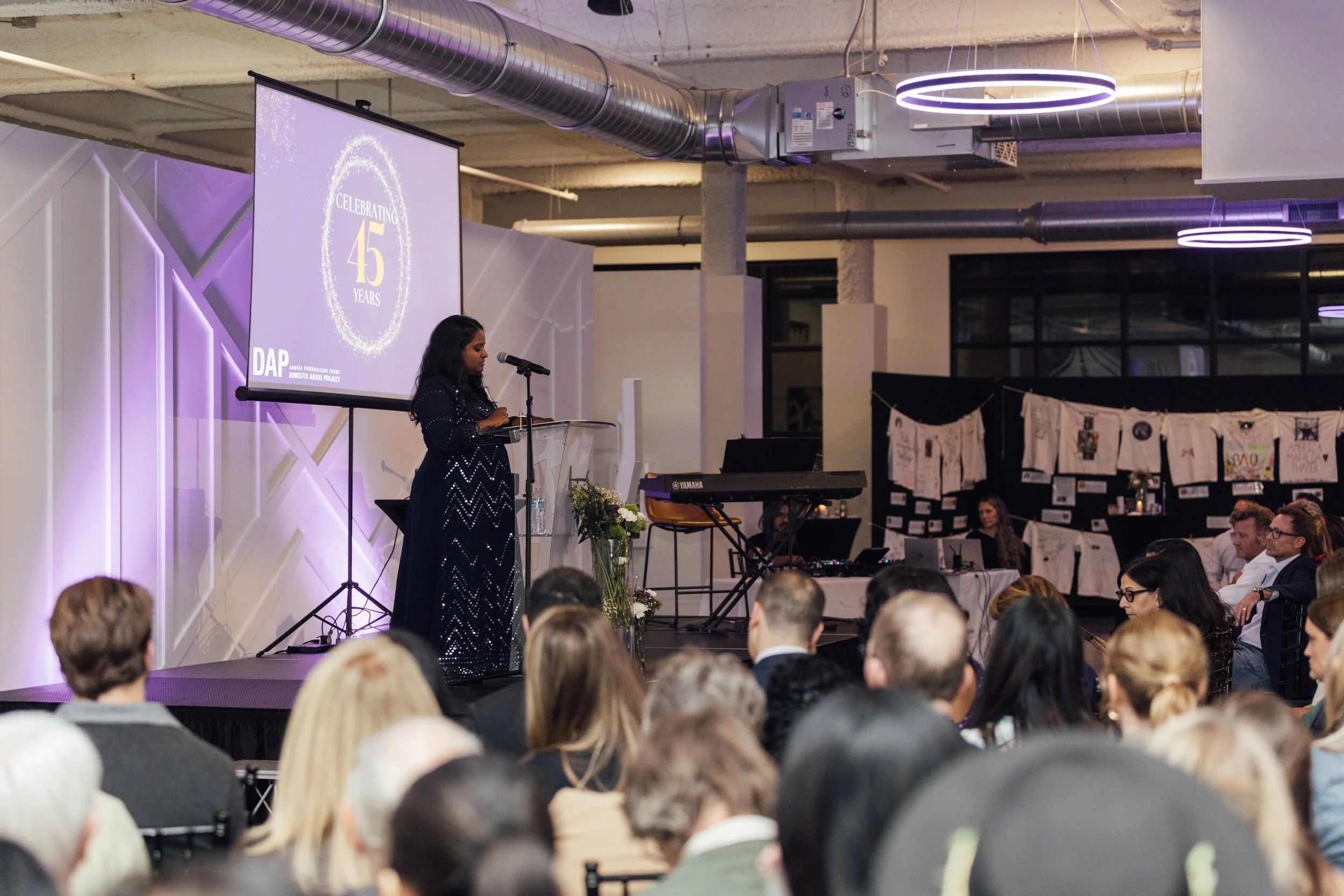 A woman giving a speech at a celebration event with a large screen behind her displaying 'Celebrating 45 Years: DAP'. The audience is seated and watching attentively.
