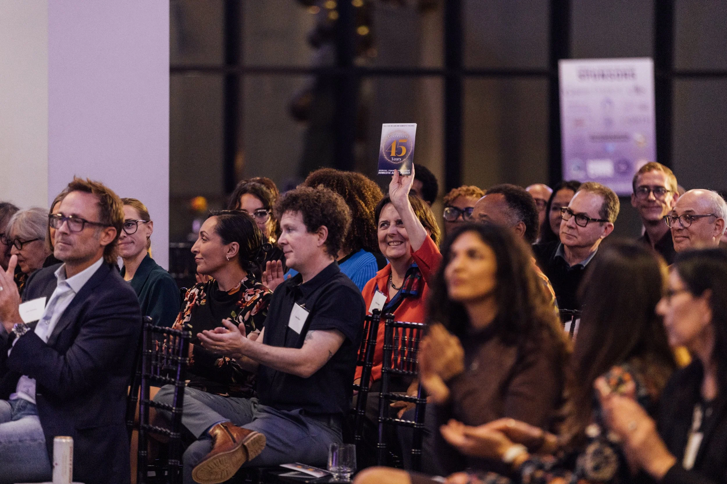 A woman in a red jacket is holding up a sign that says 'Celebrating 45 Years' at an event with a diverse audience seated and clapping.