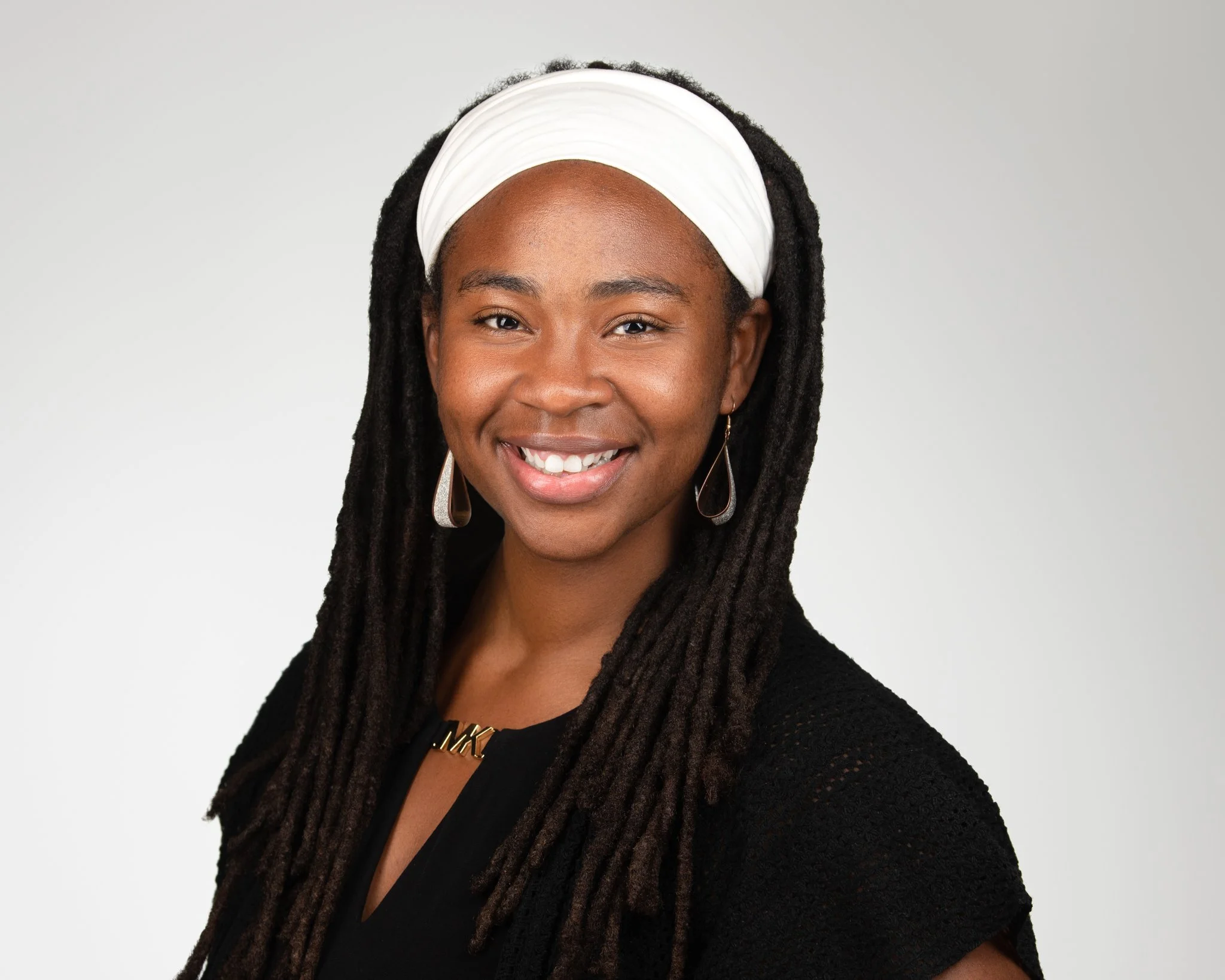 Portrait of an African American woman with long dreadlocks, wearing a white headband, black attire, and earrings, smiling at the camera against a gray background.