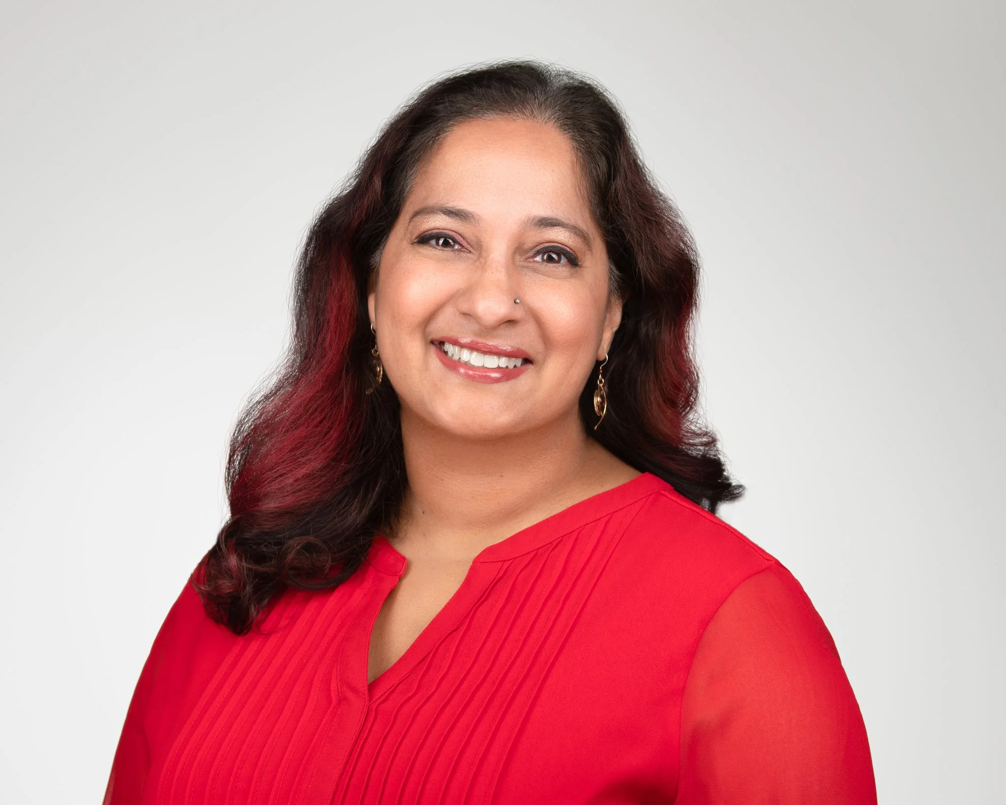 Portrait of a woman with dark hair with red highlights, smiling, wearing a red top and gold earrings, against a plain light gray background.