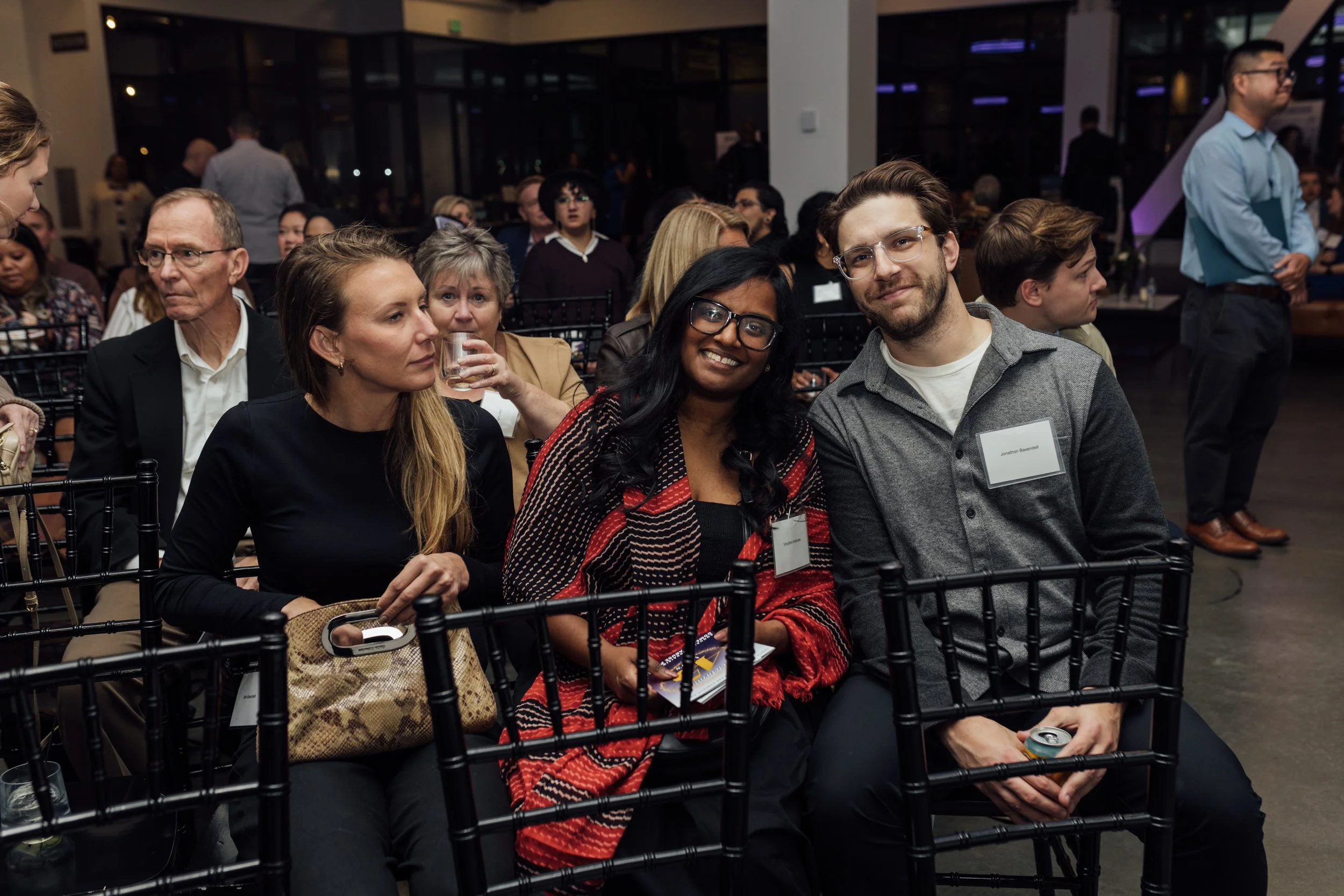 Three people sitting in the front row at a conference or event, smiling and looking at the camera, with many others seated behind them in a large, modern indoor space.