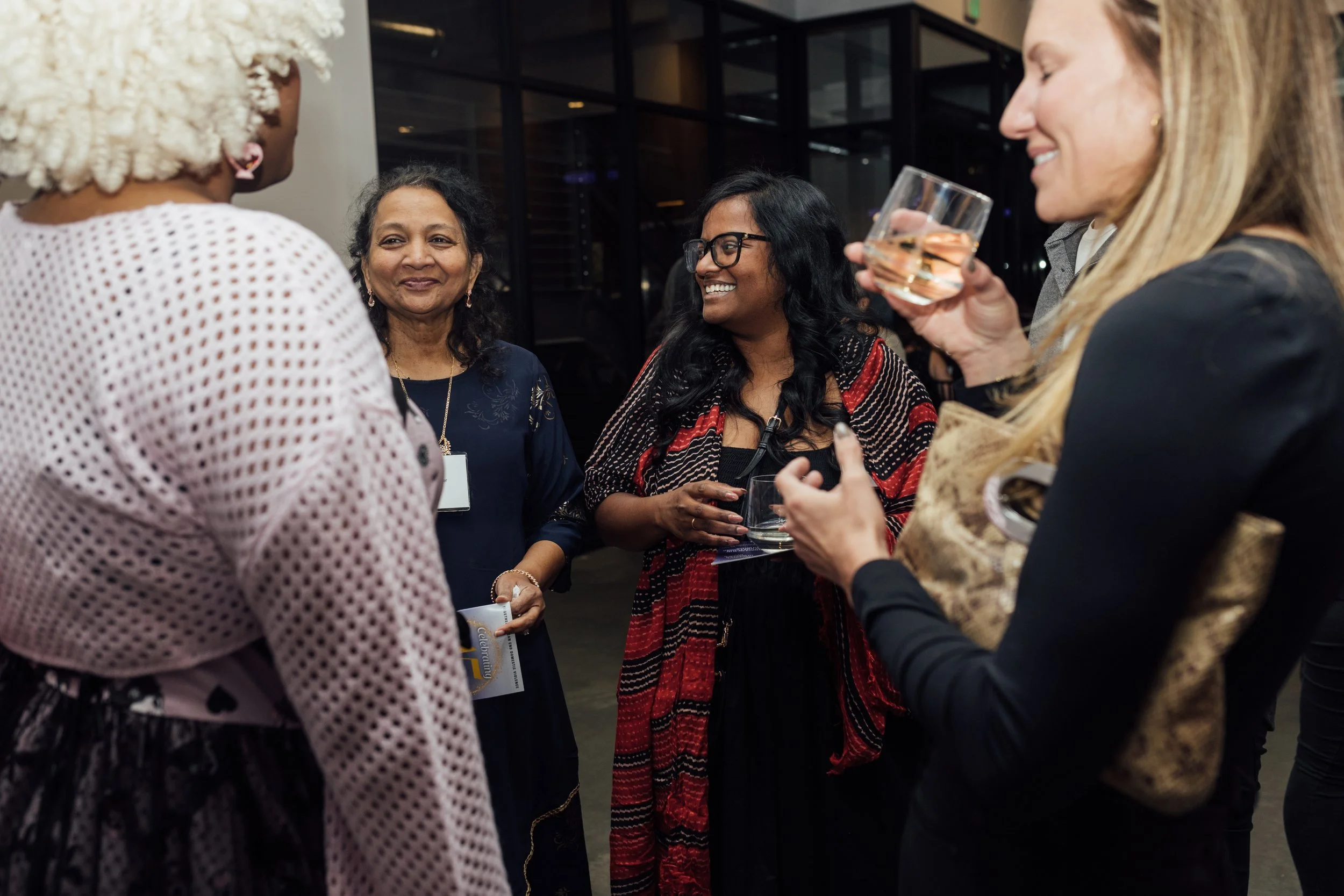 Group of four women at a social gathering, engaging in conversation, holding glasses of drinks, smiling, indoors with a modern background.