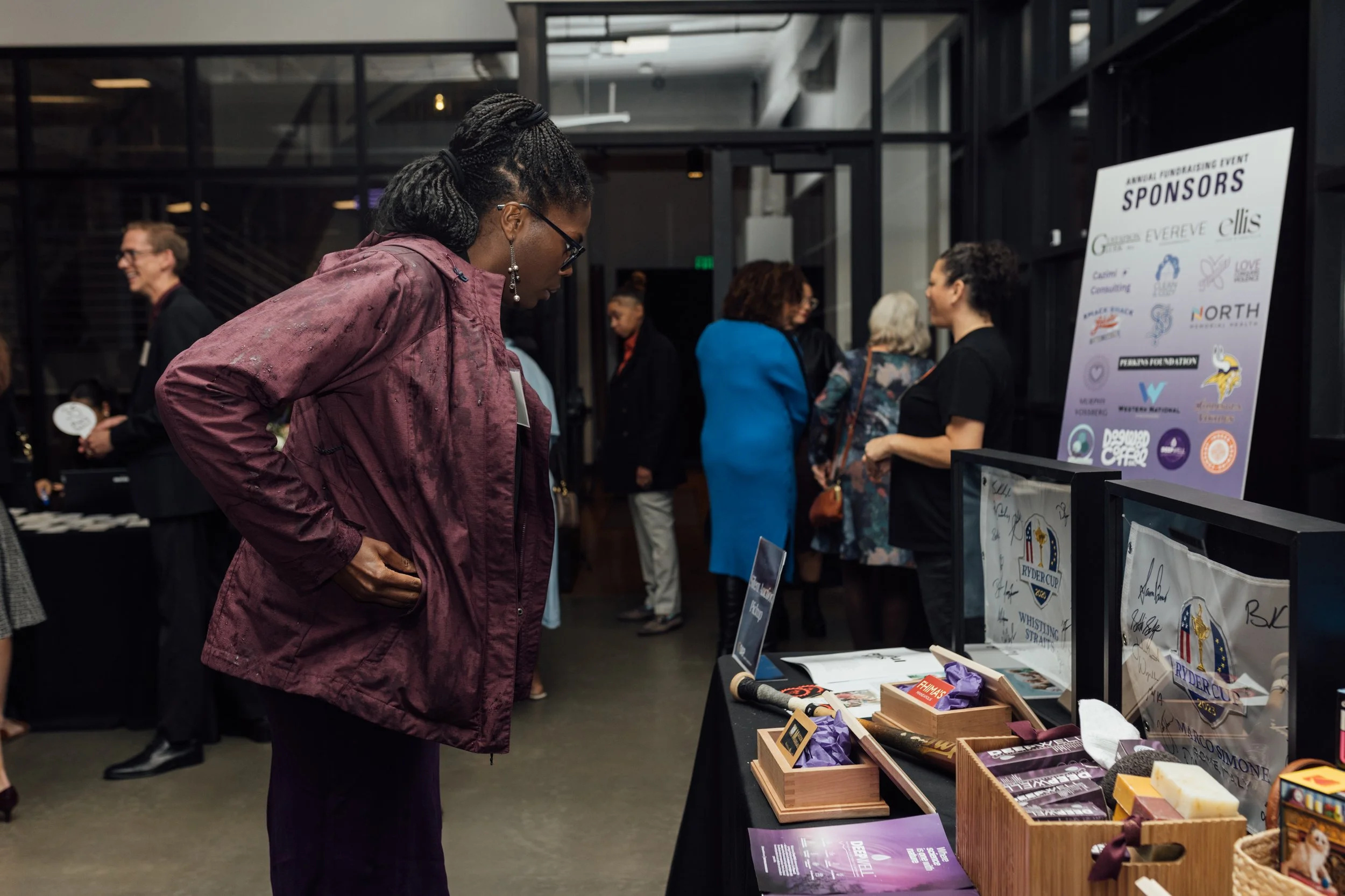 A woman in a maroon jacket and glasses looks at items displayed on a table at a fundraising event, with other attendees mingling in the background.