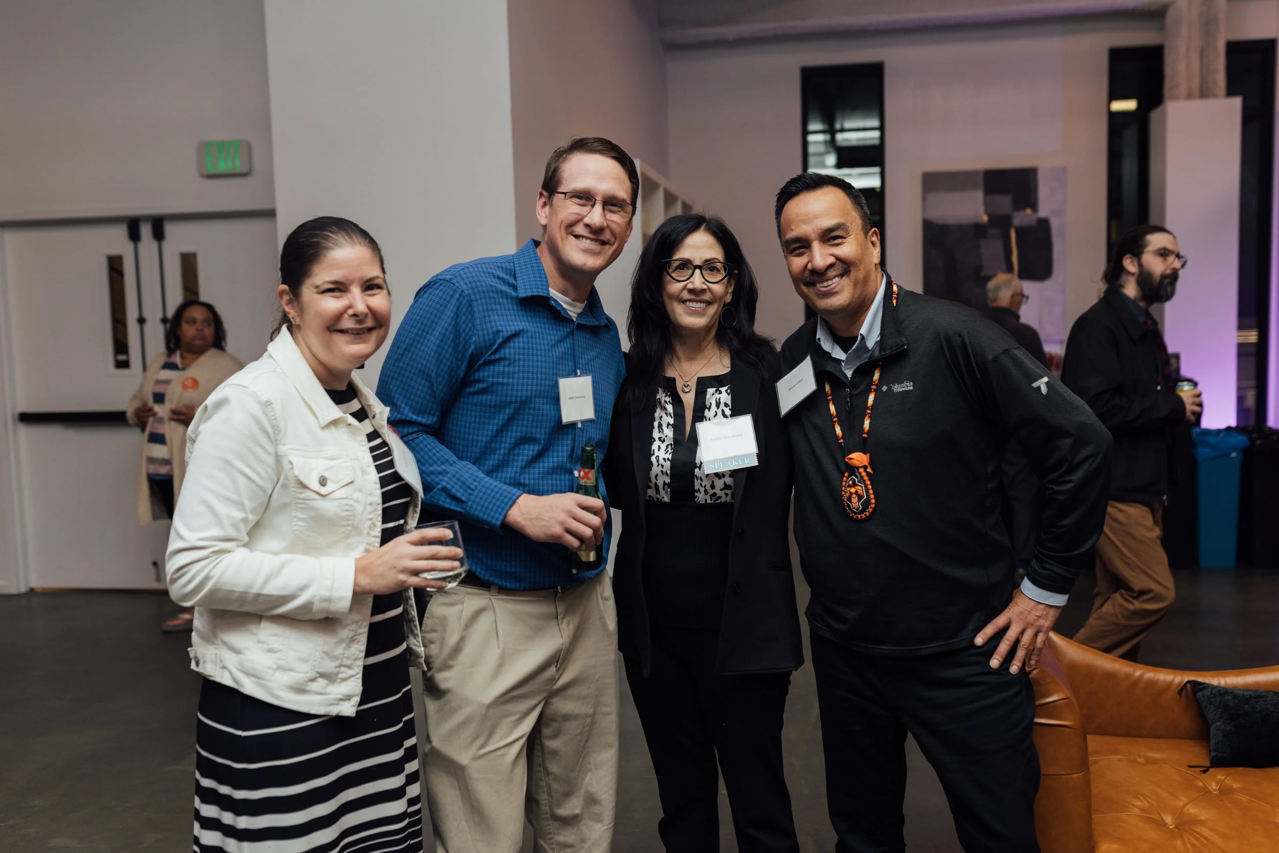 Four people smiling and posing for a photo at a social event or gathering indoors, with two men and two women.