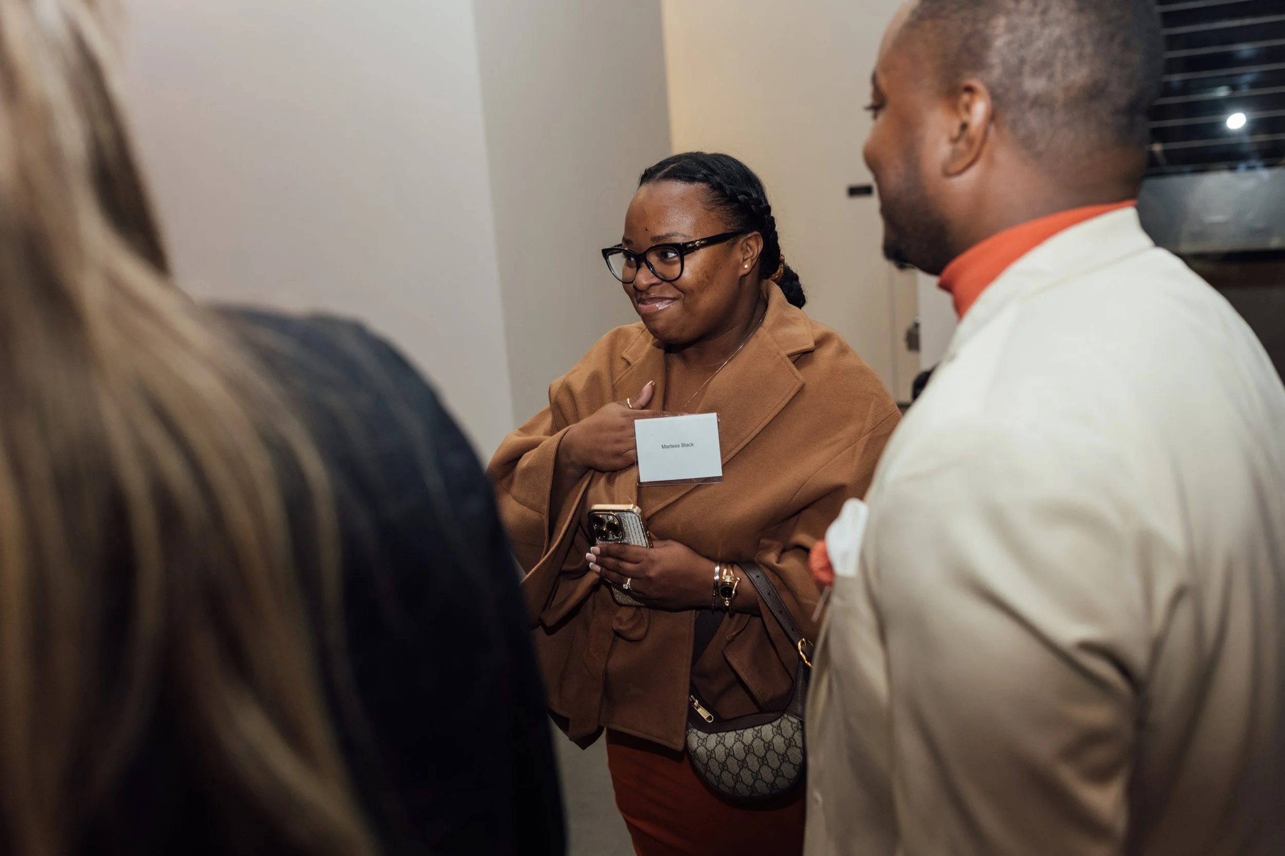 A woman wearing glasses with a name tag that says 'Martess Black' is engaged in conversation with a group of people. She is smiling and holding a phone, wearing a tan jacket and a Gucci purse.