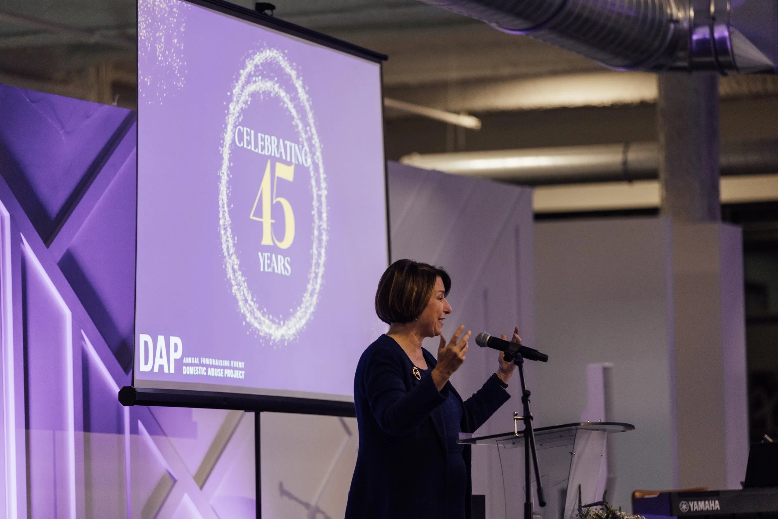 A woman speaking at a podium during a celebration event with a large screen behind her displaying a graphic that says 'Celebrating 45 years' and a logo for Domestic Abuse Project (DAP).