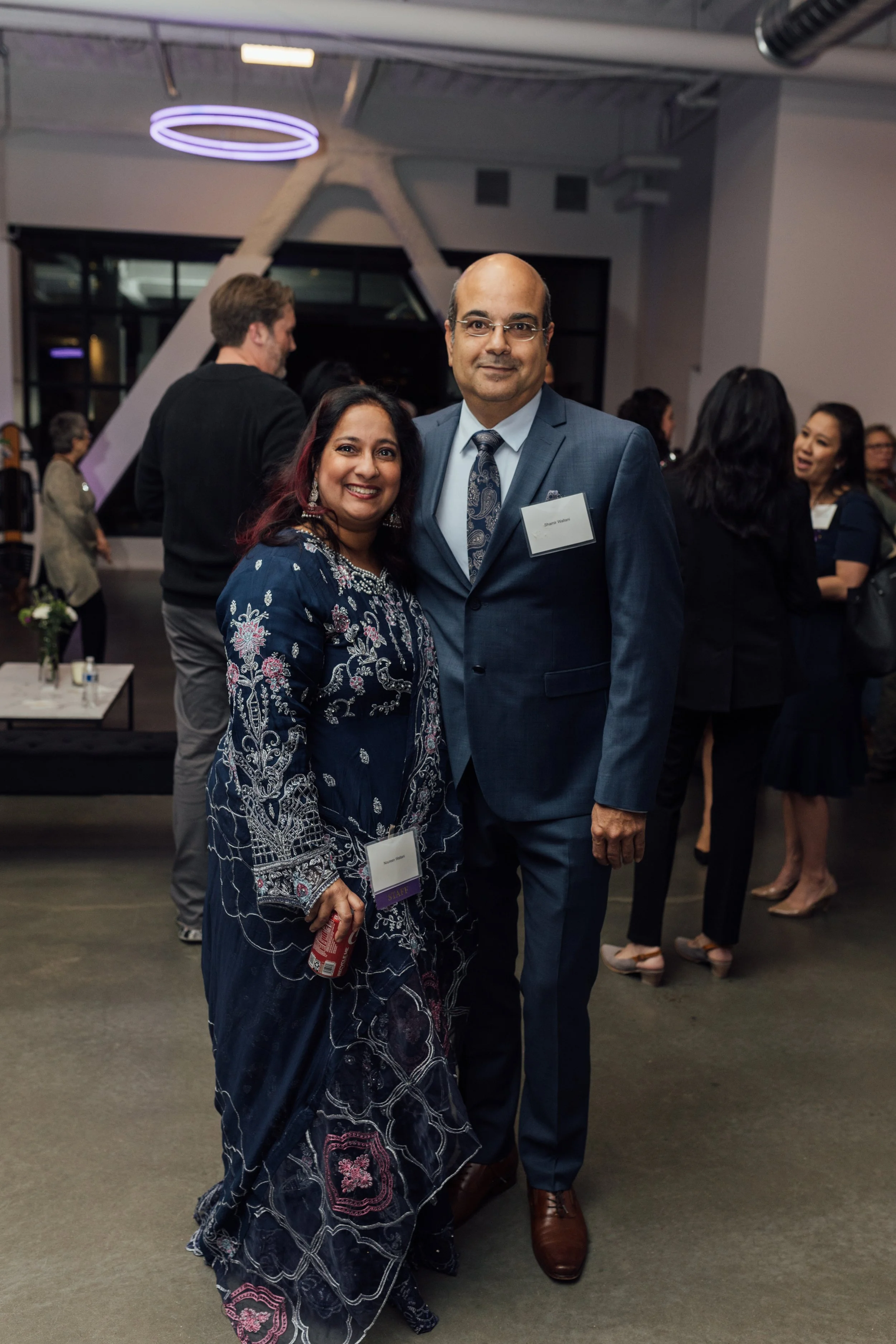 A woman and a man in formal attire posing together at an event in a modern, well-lit indoor space, with other people socializing in the background.