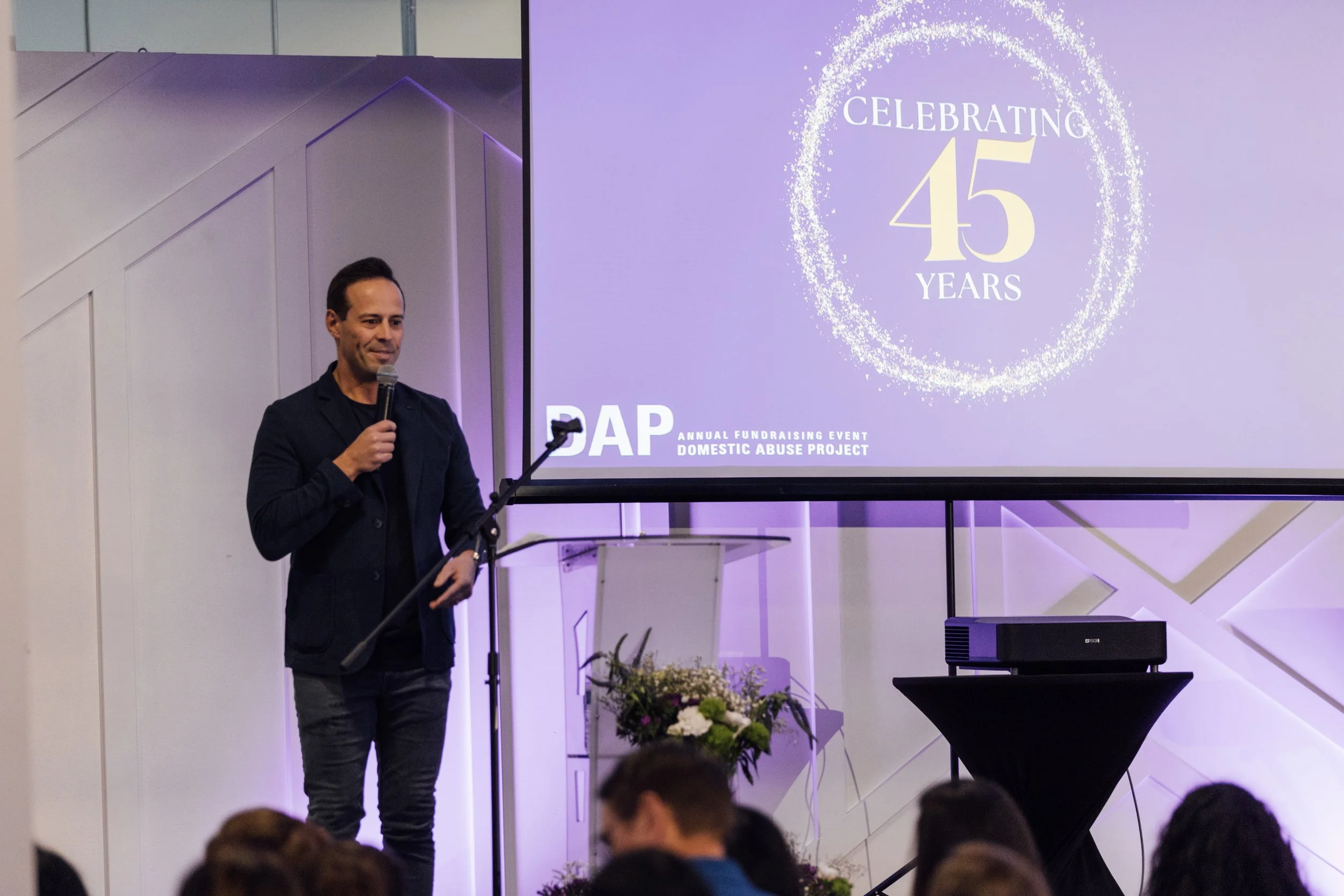 A man standing with a microphone in hand, giving a speech at an event celebrating 45 years for the Domestic Abuse Project, with a large purple screen displaying the event logo and text in the background.