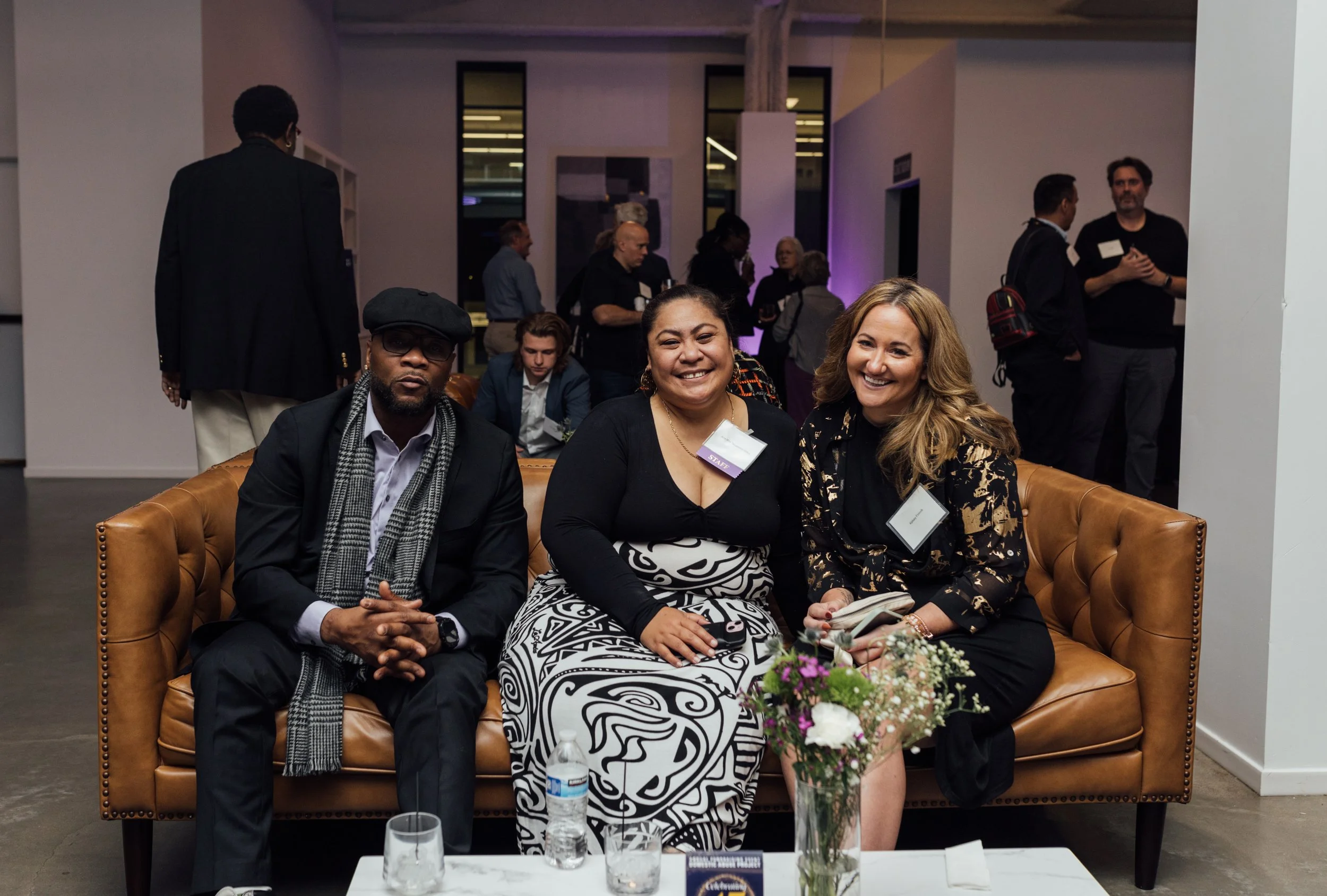 Three people sitting on a brown leather couch at a social event, smiling for the camera with a table in front holding a water bottle, glasses, and a bouquet of flowers. There are other individuals in the background engaged in conversations.