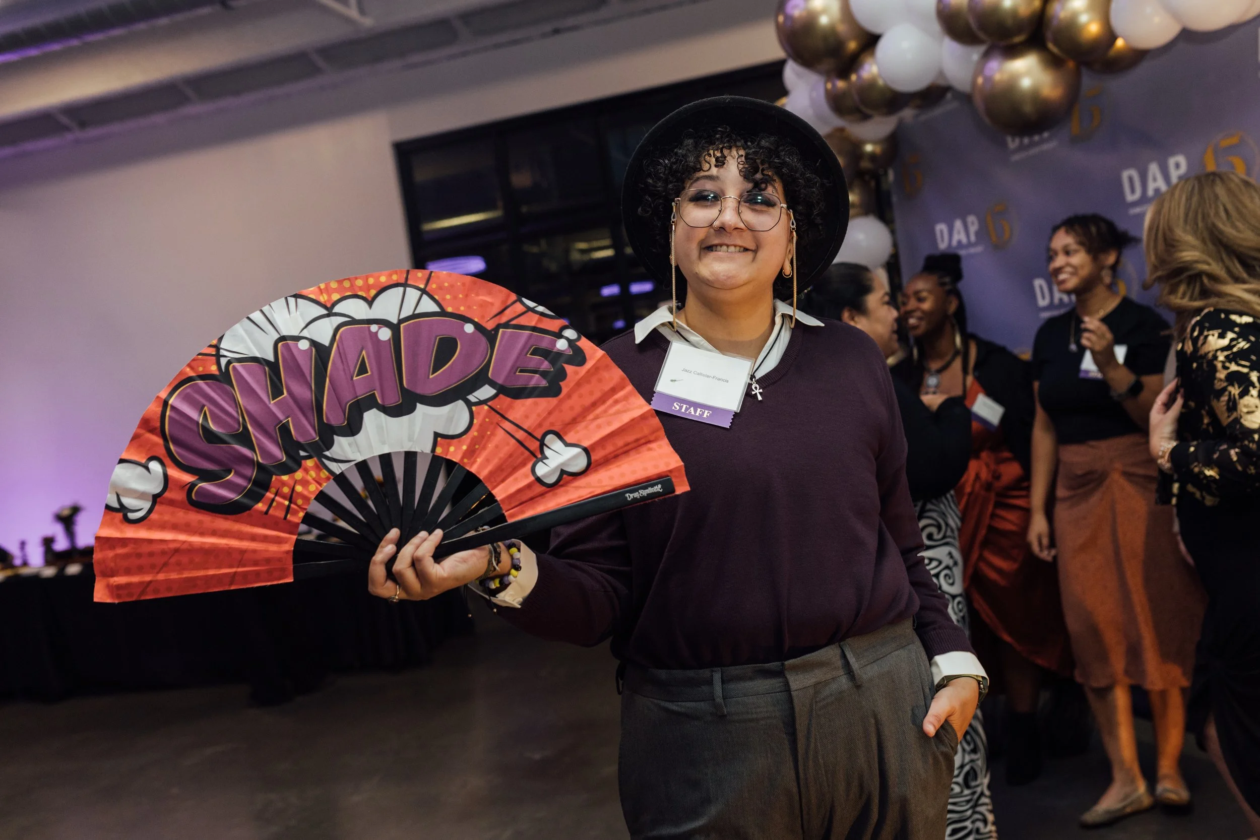 A woman holding a large fan with the word 'SHADES' written on it, smiling at a social gathering with balloons and people in the background.