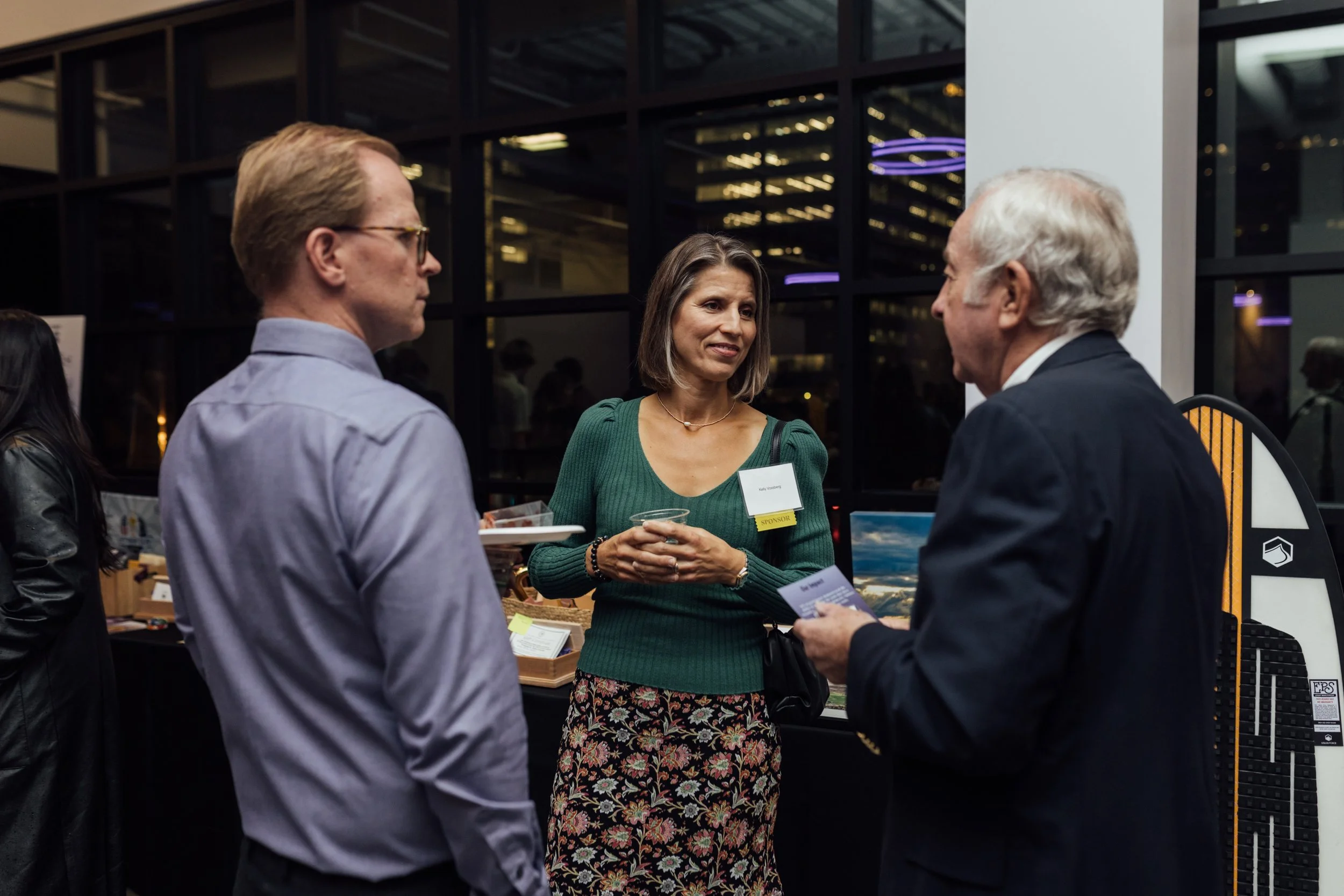 Three people engaging in conversation at an indoor networking event, with a woman holding a drink and wearing a name badge, and two men listening attentively.