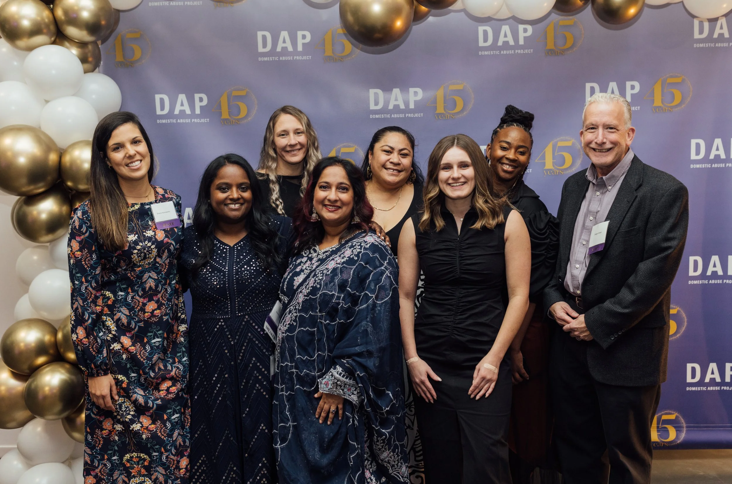 A group of nine people, women and men, standing together and smiling at an event with a blue background that reads 'DAP 45 Years' and 'Domestic Abuse Project.' There are gold and white balloons on the side.