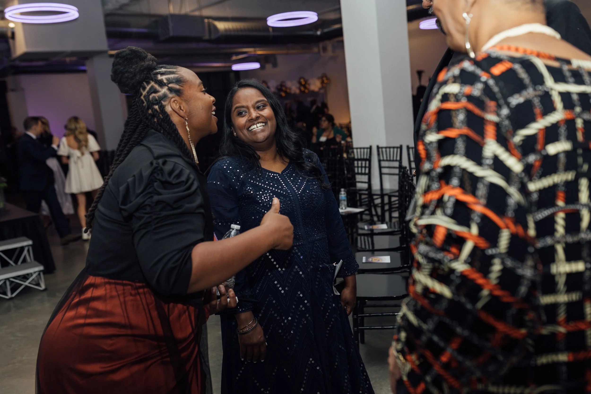 Three women engaged in conversation at a social event in a decorated venue, with empty chairs in the background.