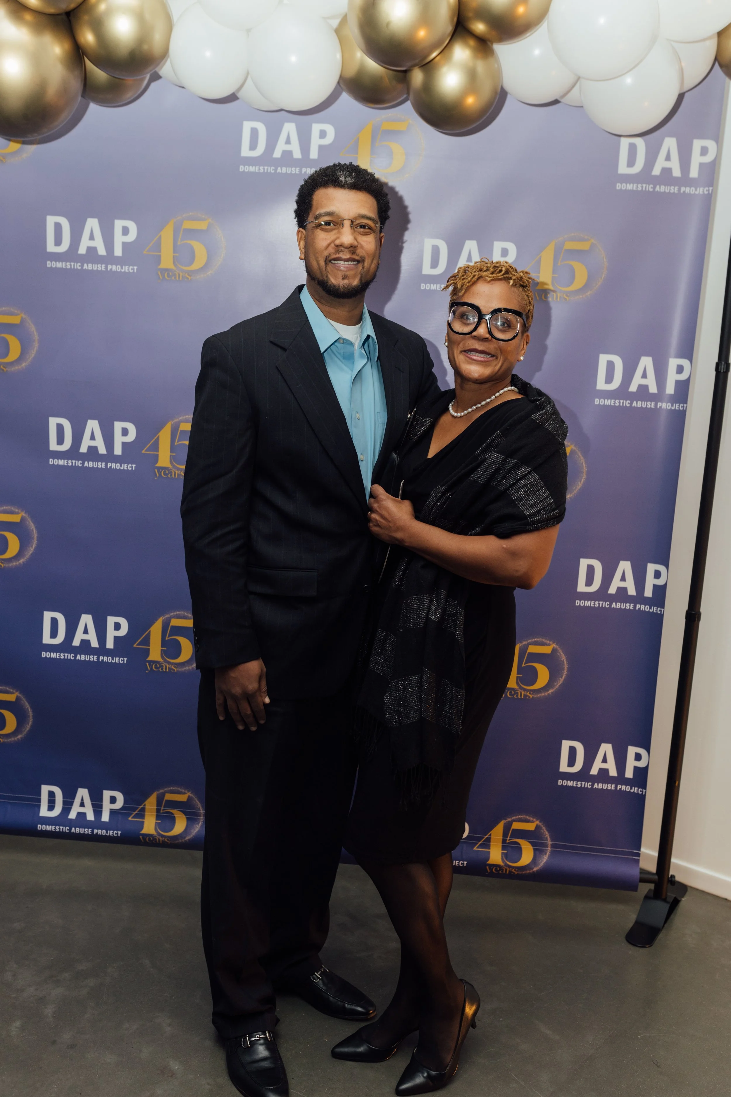 A man and woman standing together at a Domestic Abuse Project 45th anniversary event, posing in front of a backdrop with balloons. The man is wearing a black suit and blue shirt, and the woman is dressed in black with a shawl, heels, and glasses.