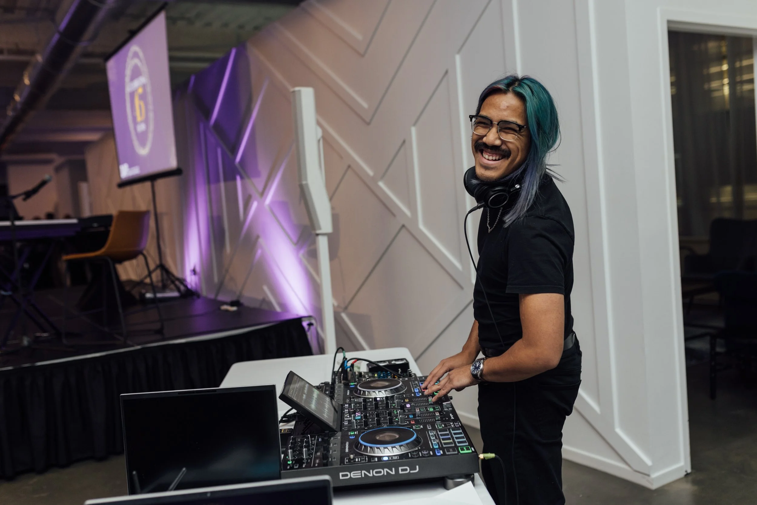 A DJ with teal and blue hair smiling and wearing glasses, standing behind a DJ console with headphones around his neck, in a modern indoor venue with stage and purple lighting.