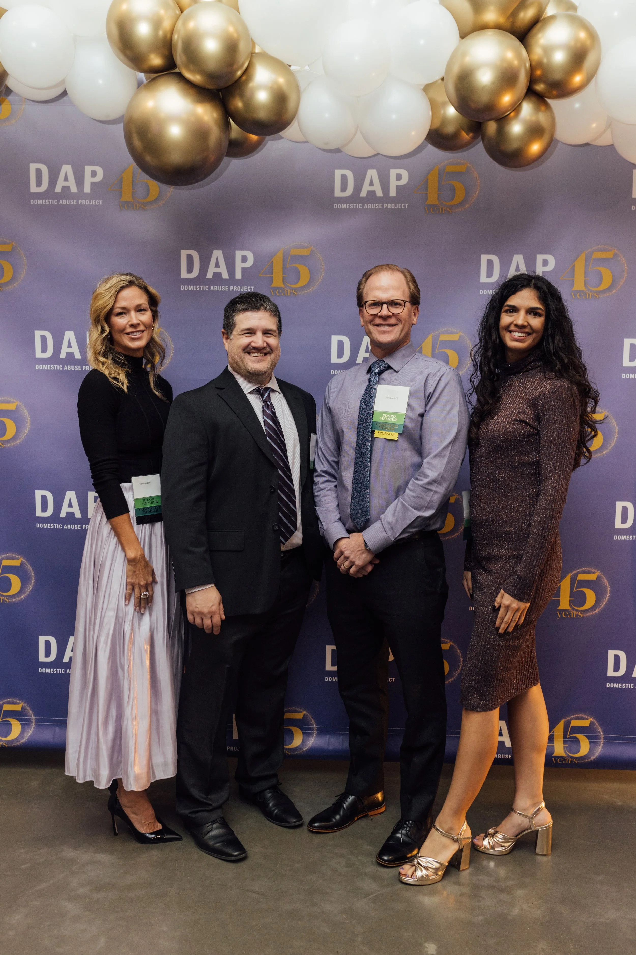 Four people dressed in formal attire standing in front of a DAP 45th anniversary backdrop with gold and white balloons overhead.