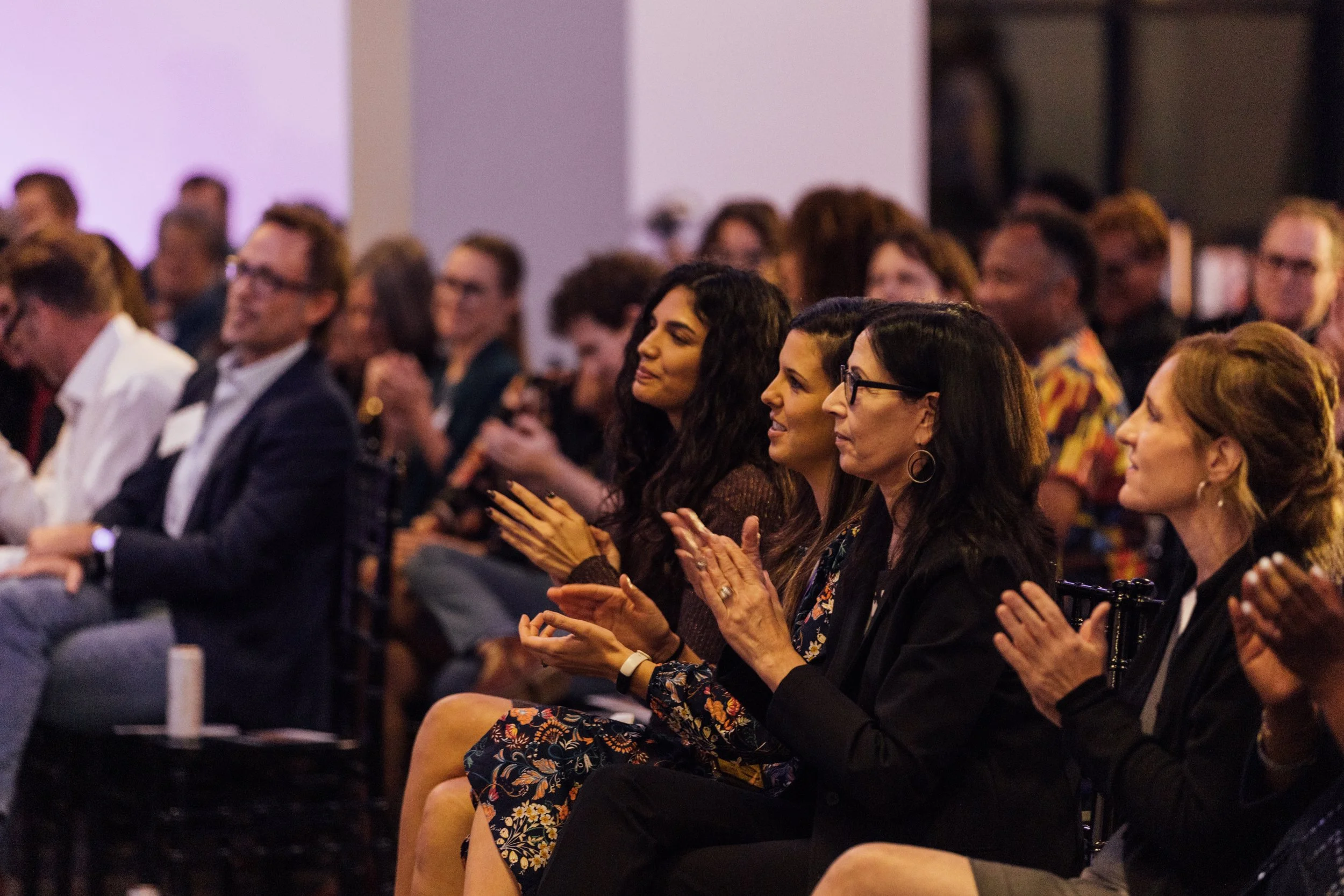 Audience of diverse professionals sitting and clapping at conference or seminar, attentively listening to speaker.