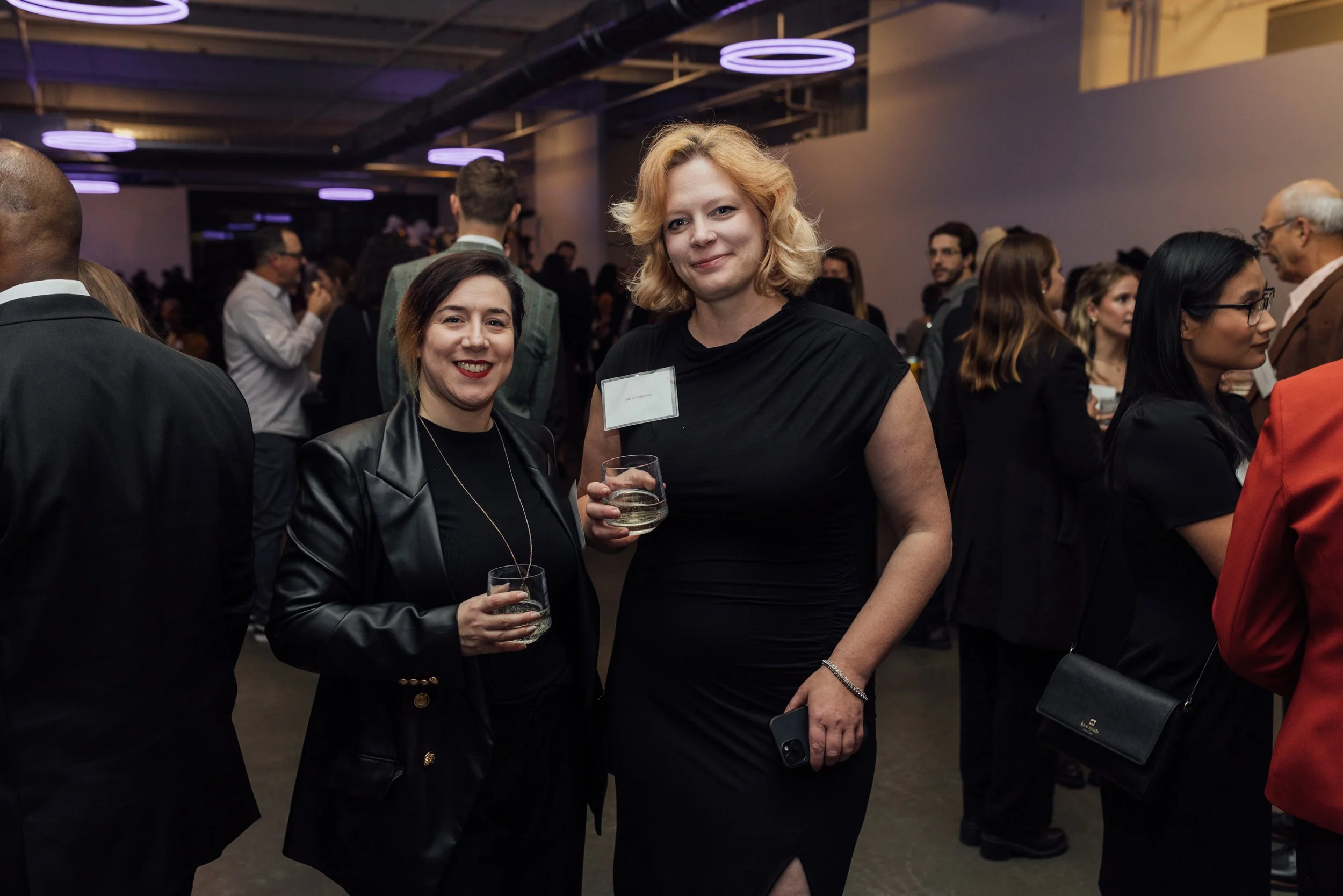 Two women in black attire holding glasses of sparkling wine at a professional networking event or reception, with a crowd of people in the background.