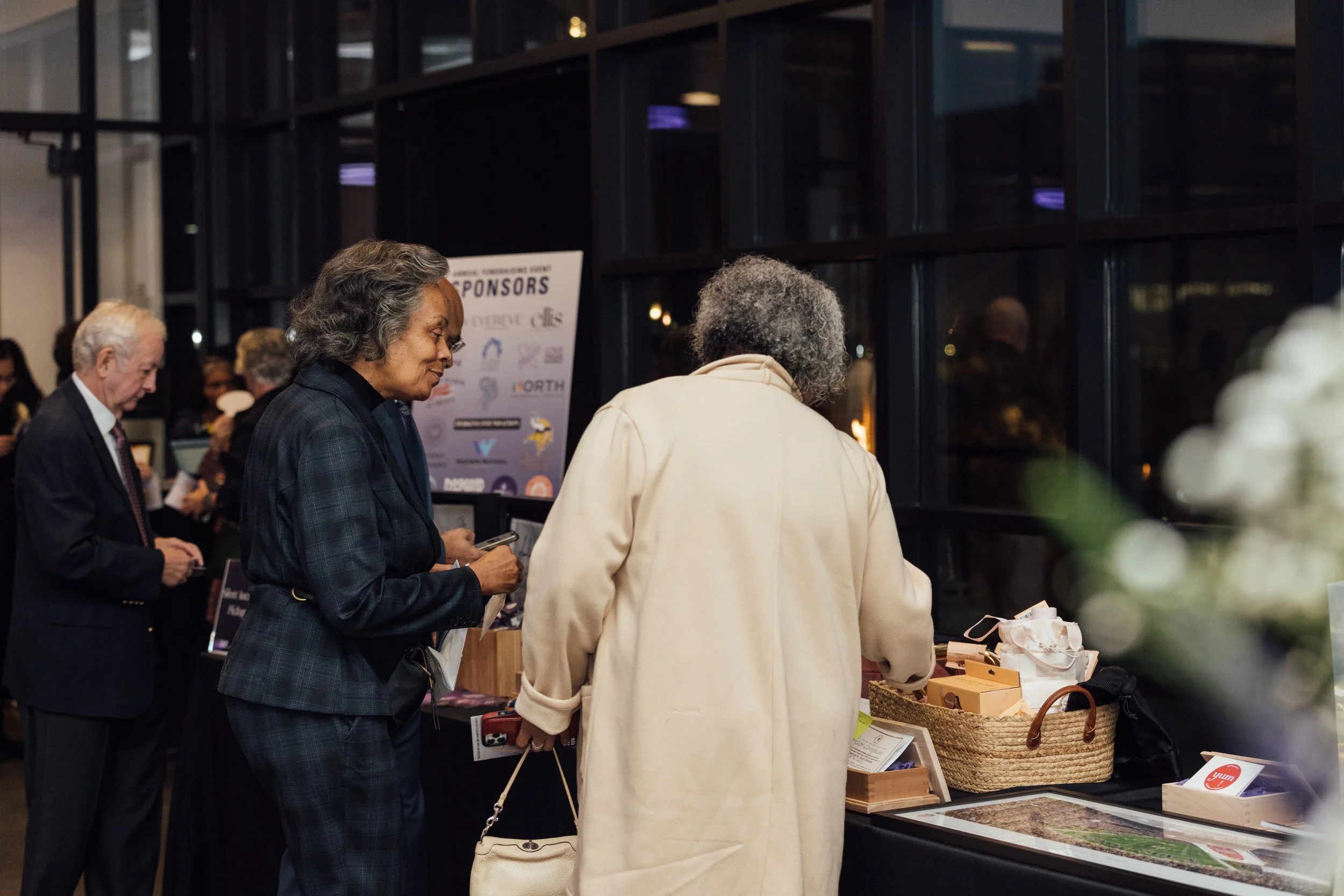 People shopping at a booth with various items at an indoor event, against a windowed wall with a poster in the background.