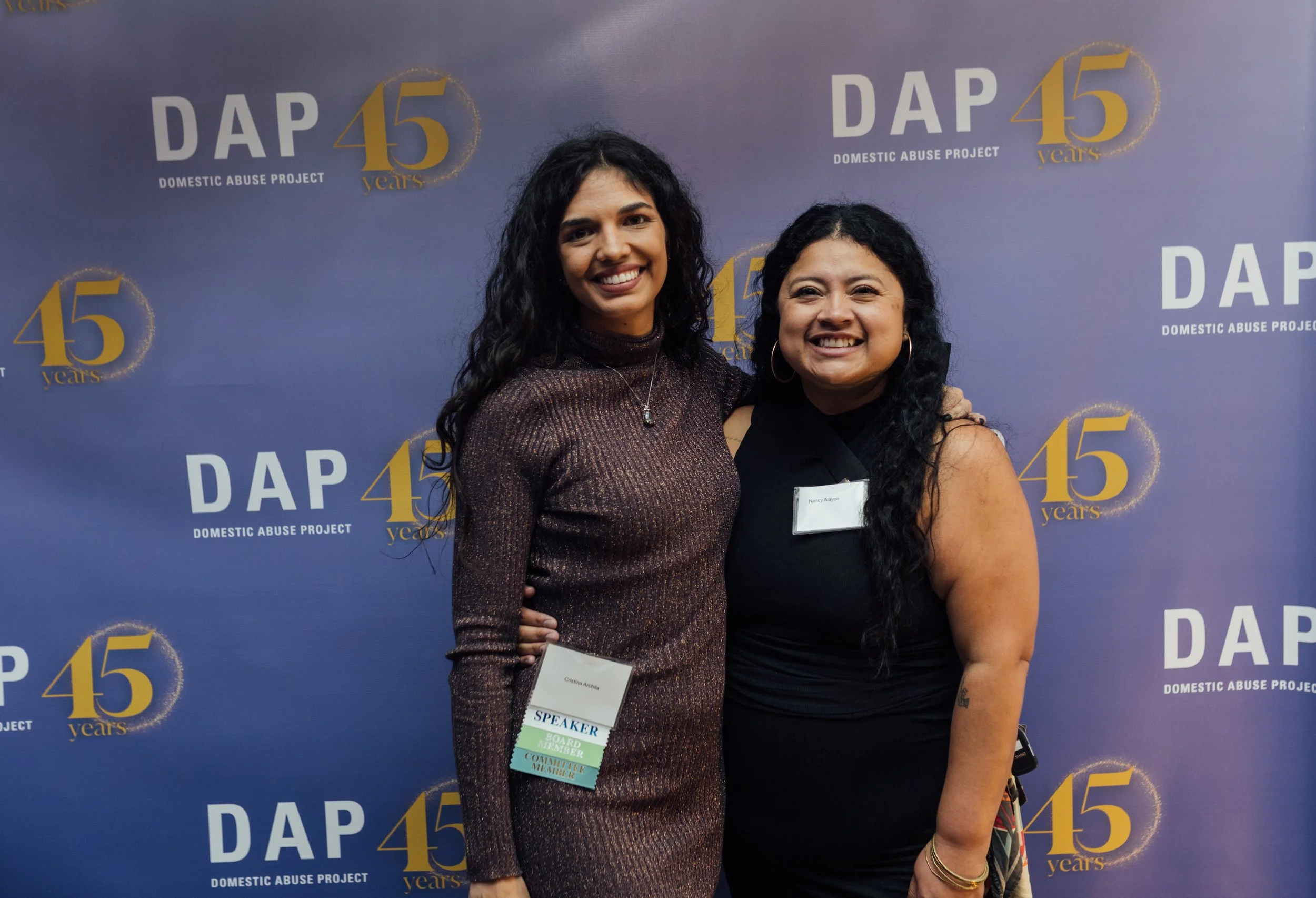 Two women smiling and standing close together at a Domestic Abuse Project event with a blue backdrop that says DAP 45 years.