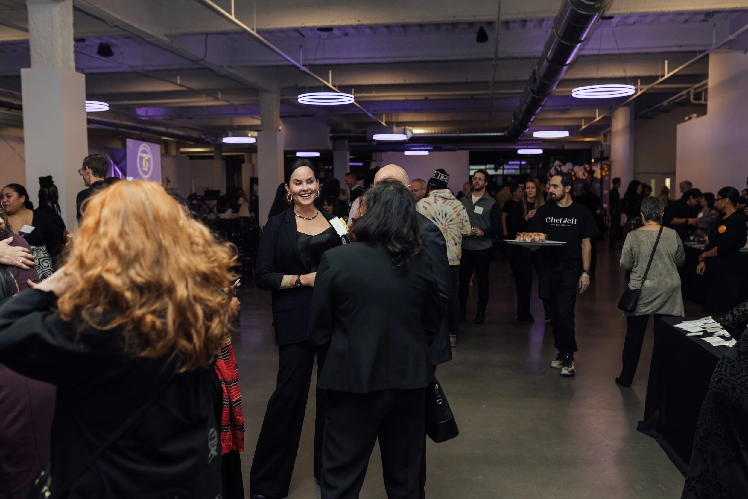 A group of people gathering at an indoor social event or mixer, engaging in conversations, with purple lighting and a modern industrial interior