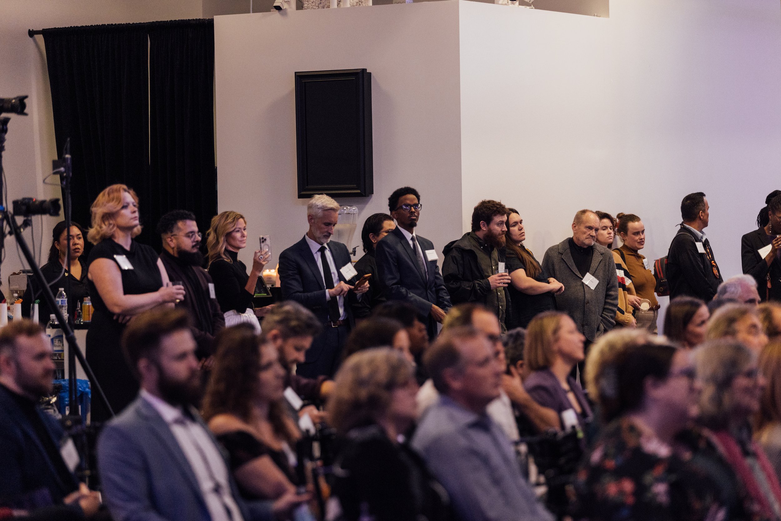 Audience members standing and sitting in a large room at a conference or event, with some looking attentively and others taking photos or videos.