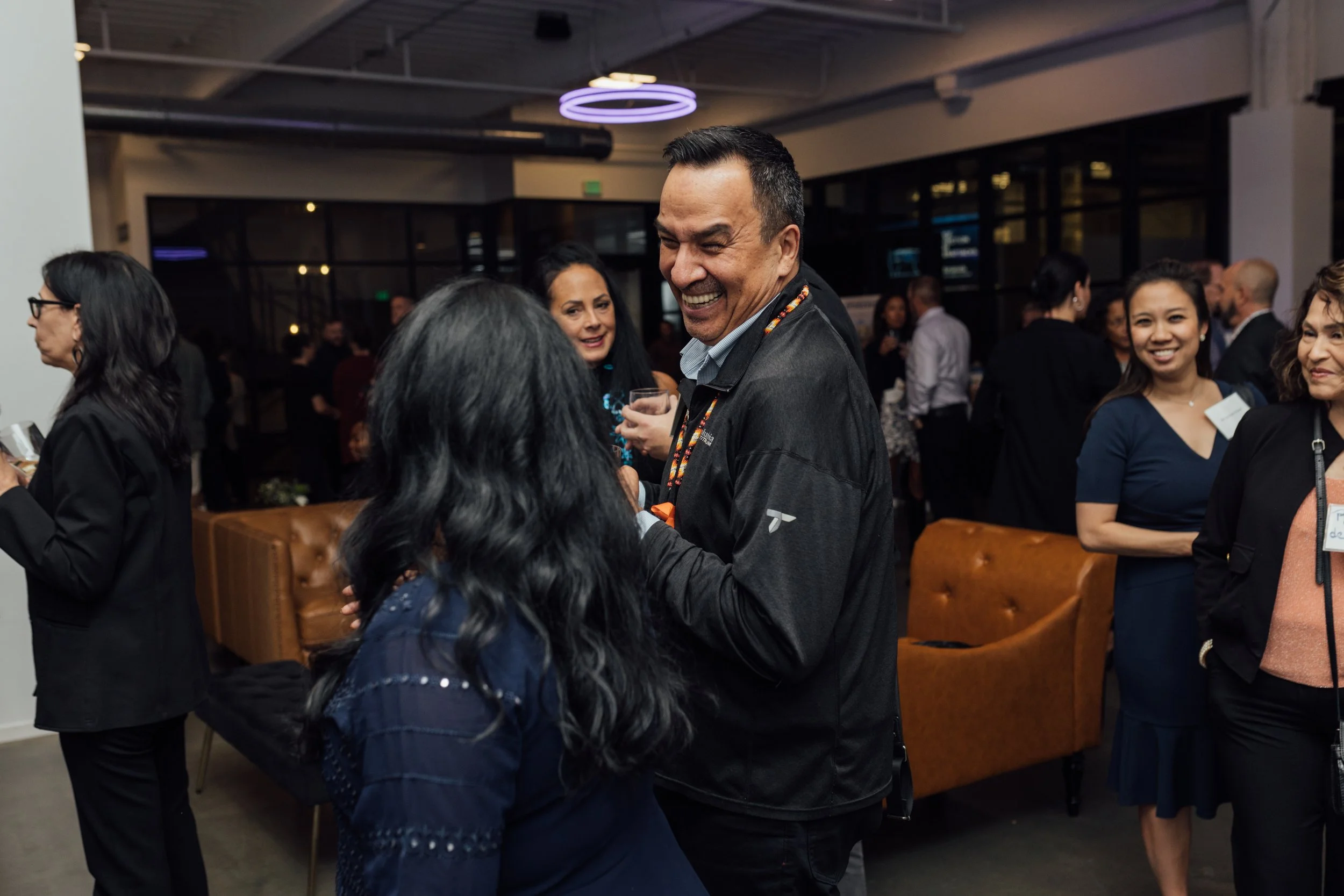People socializing at an indoor event, smiling and talking, with a focus on a man in a black jacket laughing and conversing with a woman with long black hair.