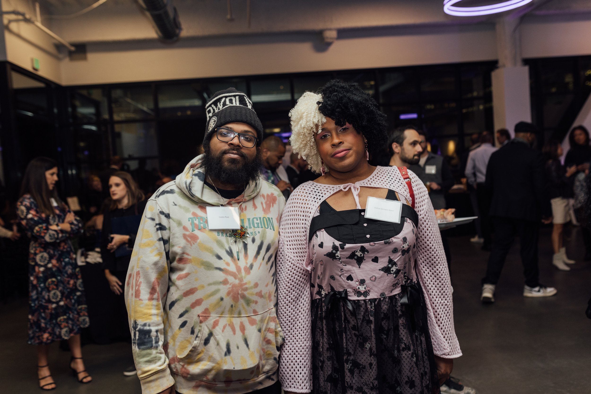 Two individuals posing together at a social event, with one wearing a tie-dye hoodie and the other wearing a pink and black dress with a black lace overlay, surrounded by other attendees in a dimly lit indoor space.
