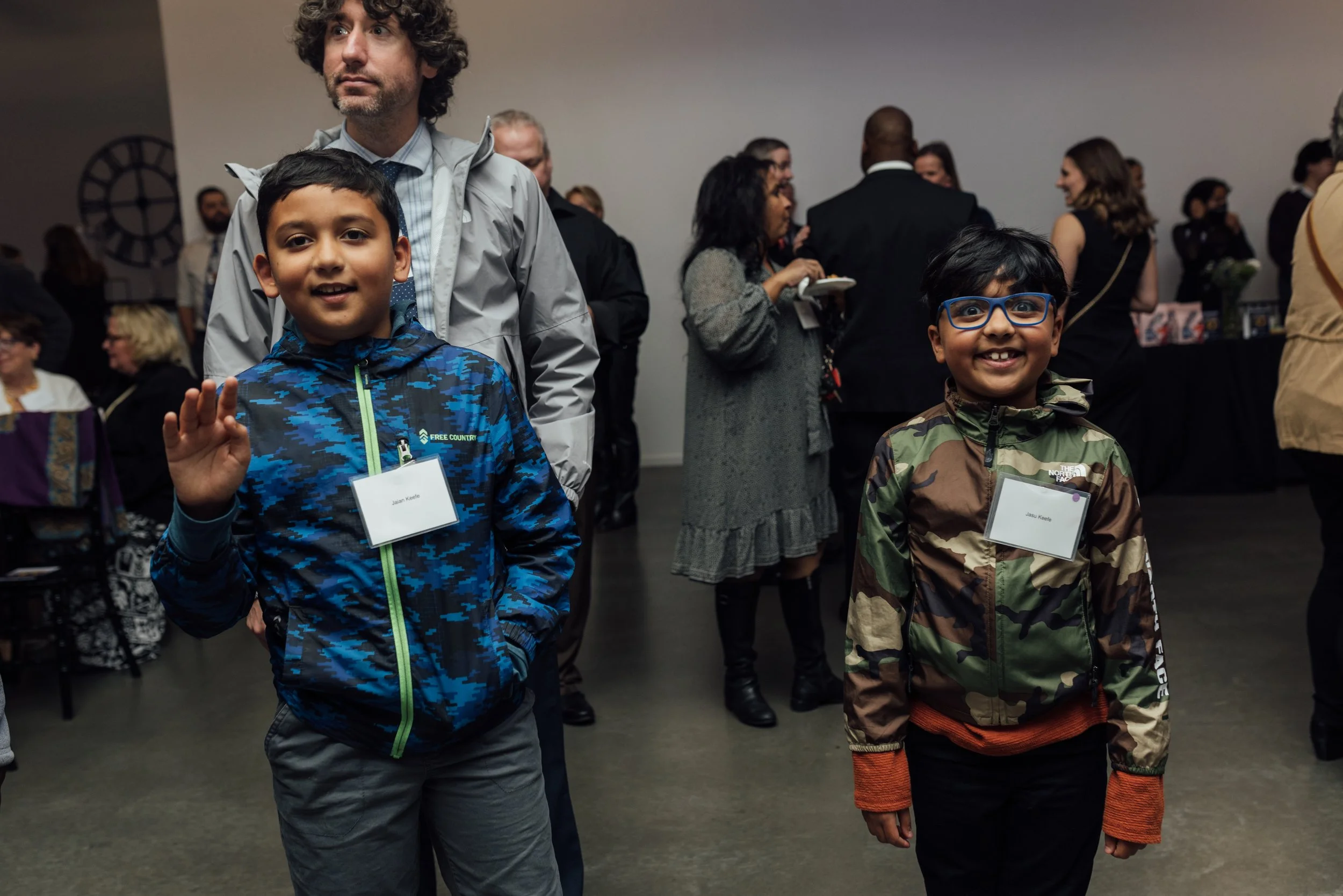 Two boys wearing jackets with name tags, one waving, standing in a crowded indoor event. One man is behind them and there are multiple people talking and mingling in the background.