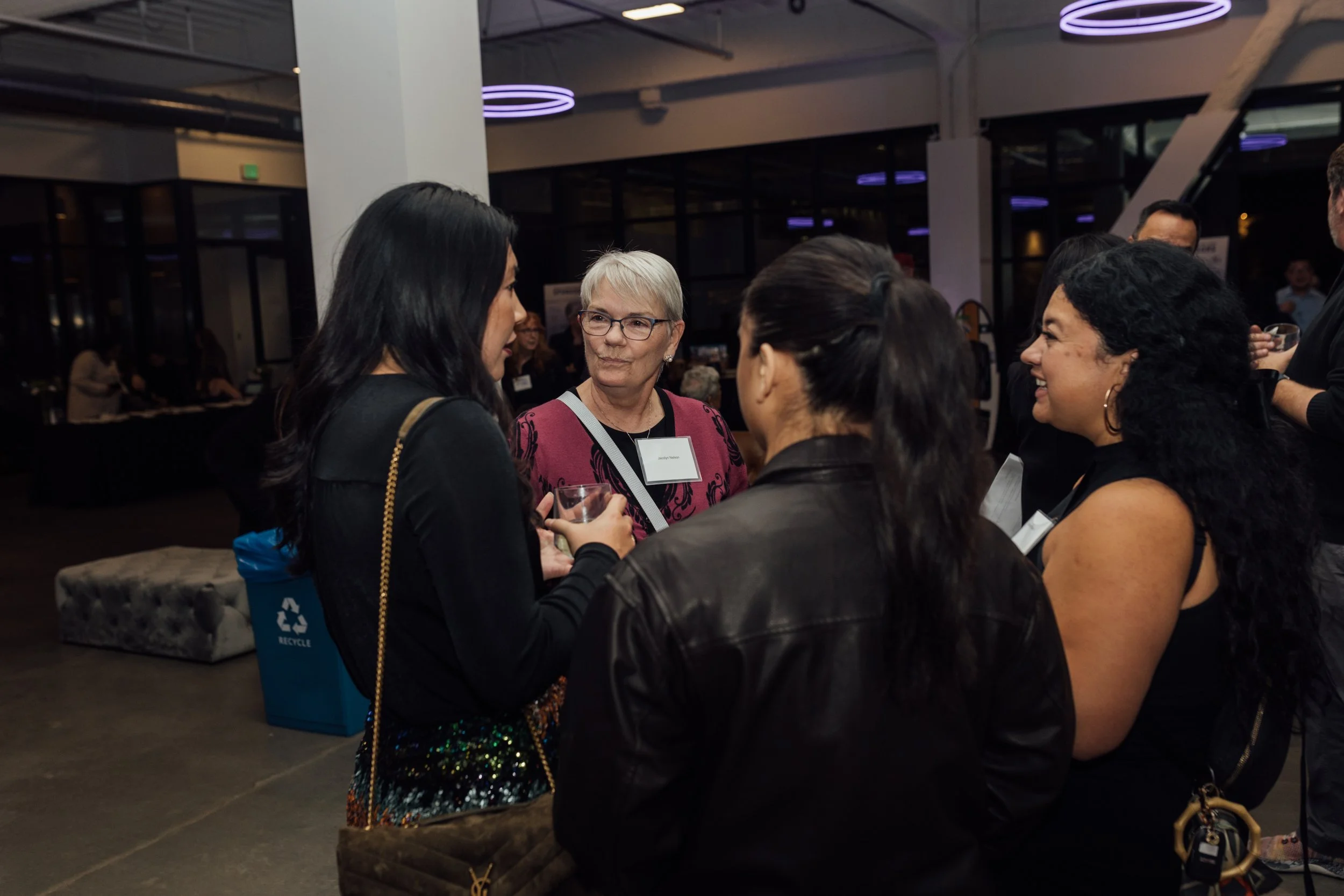 A group of women engaged in conversation at a social or professional event in a modern indoor space with purple lighting.