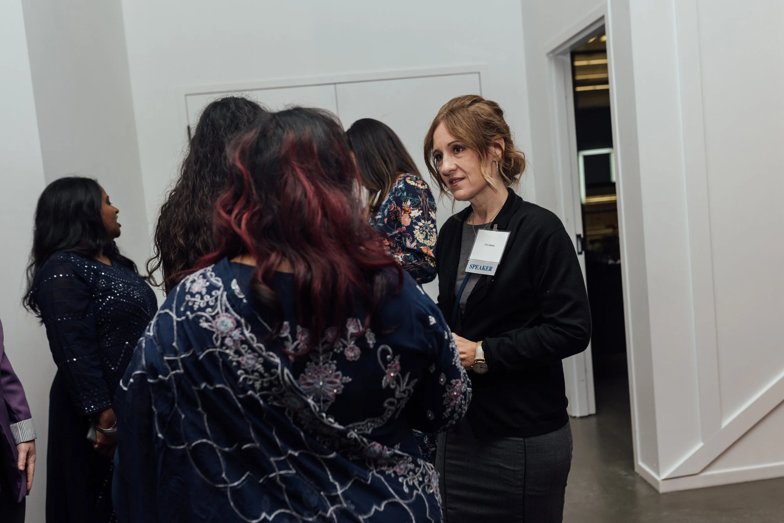 A woman with a speaker badge talking to a small group of diverse women at a conference or event in a modern, white-walled room.