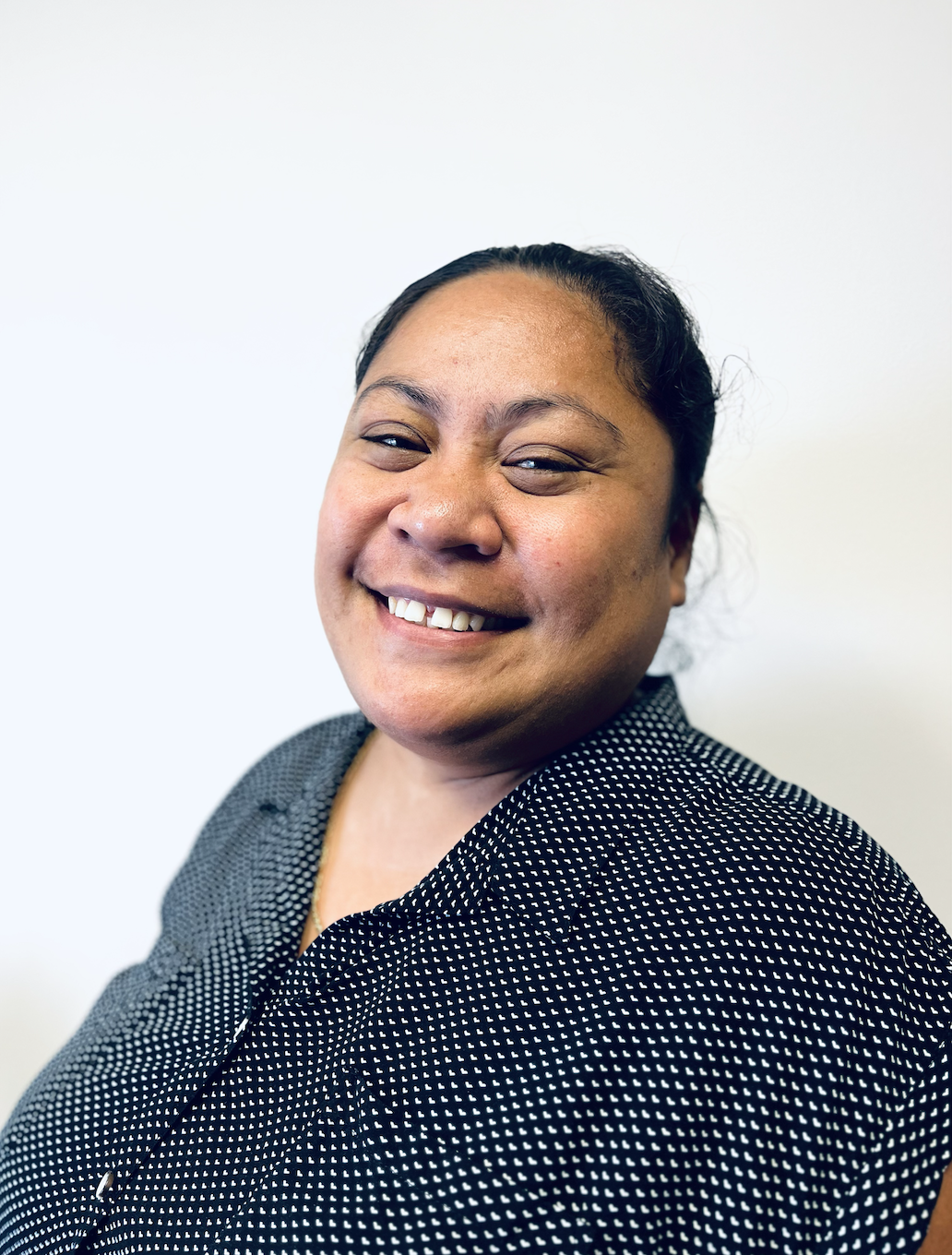 Close-up portrait of a woman with a big smile, wearing a black and white dotted shirt, standing against a plain white background.