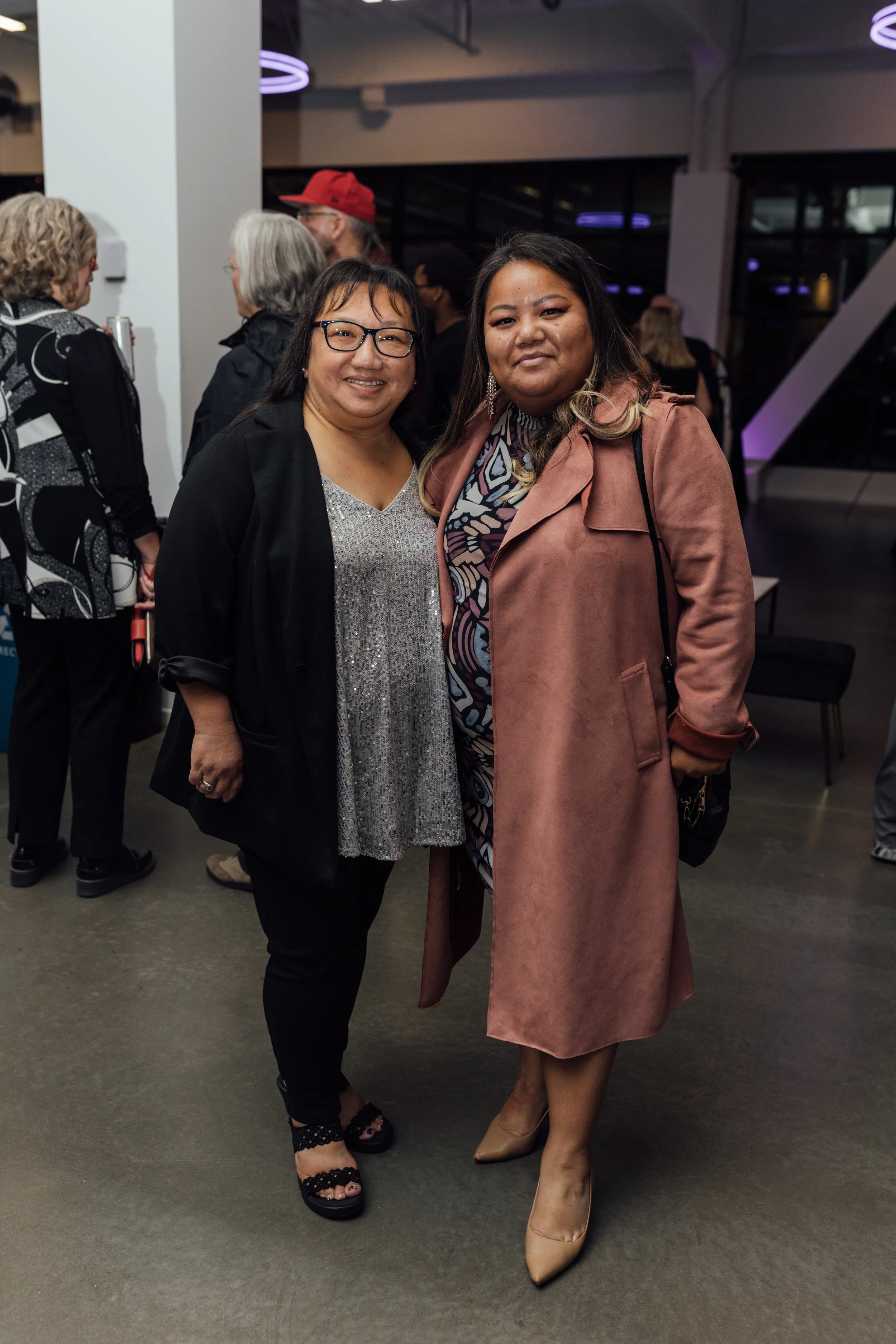 Two women standing close together at an indoor social event, smiling at the camera, with other people in the background.