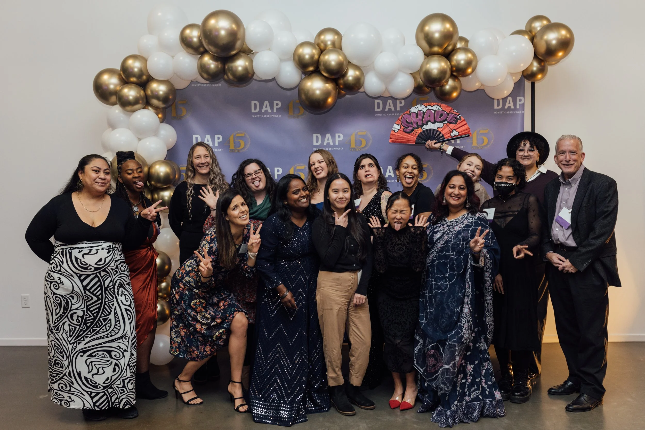 Group of diverse women and men smiling and making peace signs at a celebration with a balloon arch and a backdrop that says DAP 15