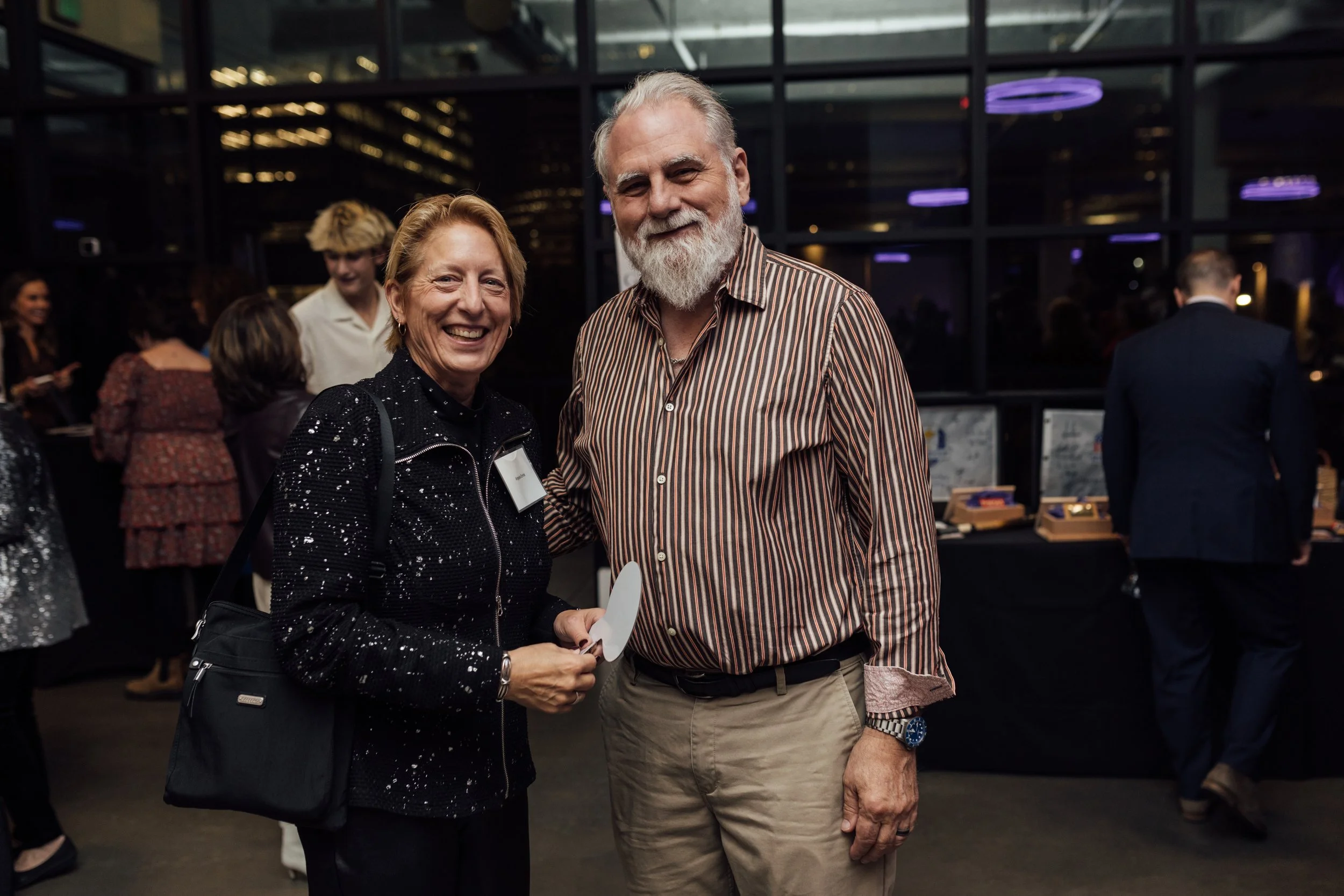 A woman and a man posing together at an indoor social event, smiling, with other people mingling in the background.