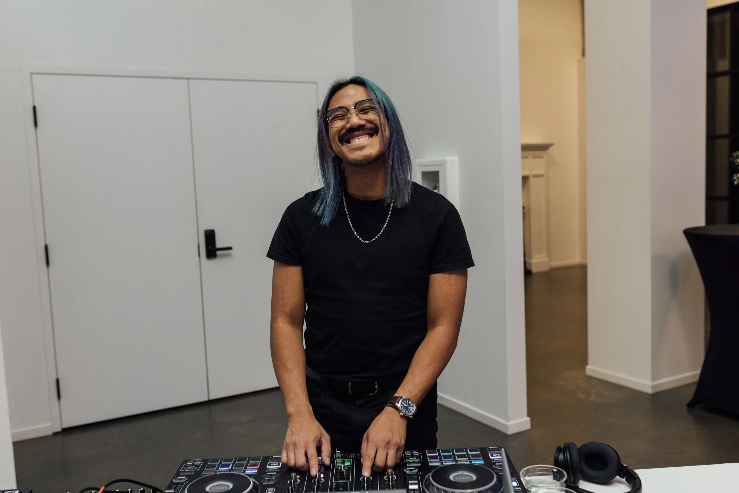 A smiling person with colorful hair and glasses standing behind DJ equipment, wearing a black t-shirt and a watch.