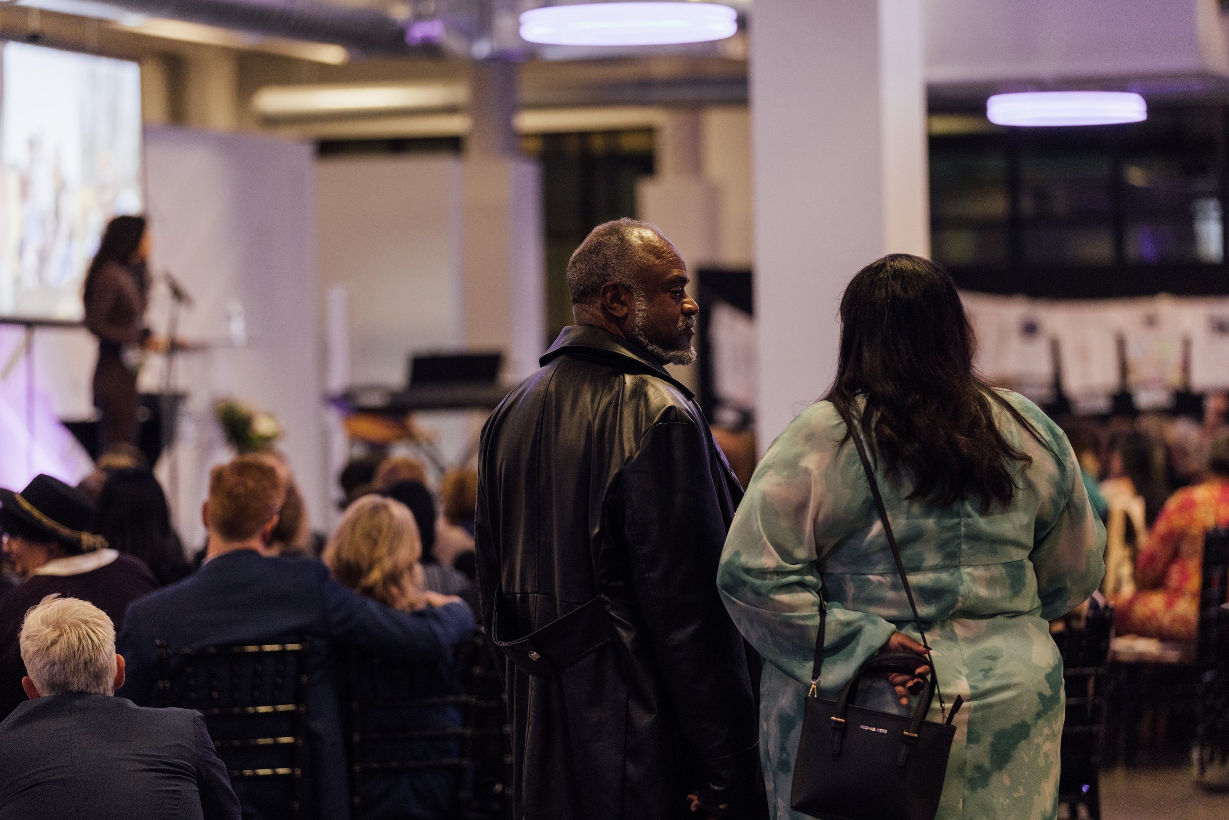 Two people, a man and a woman, standing and talking at an indoor event with a crowd seated around them. A woman is speaking on stage in the background.
