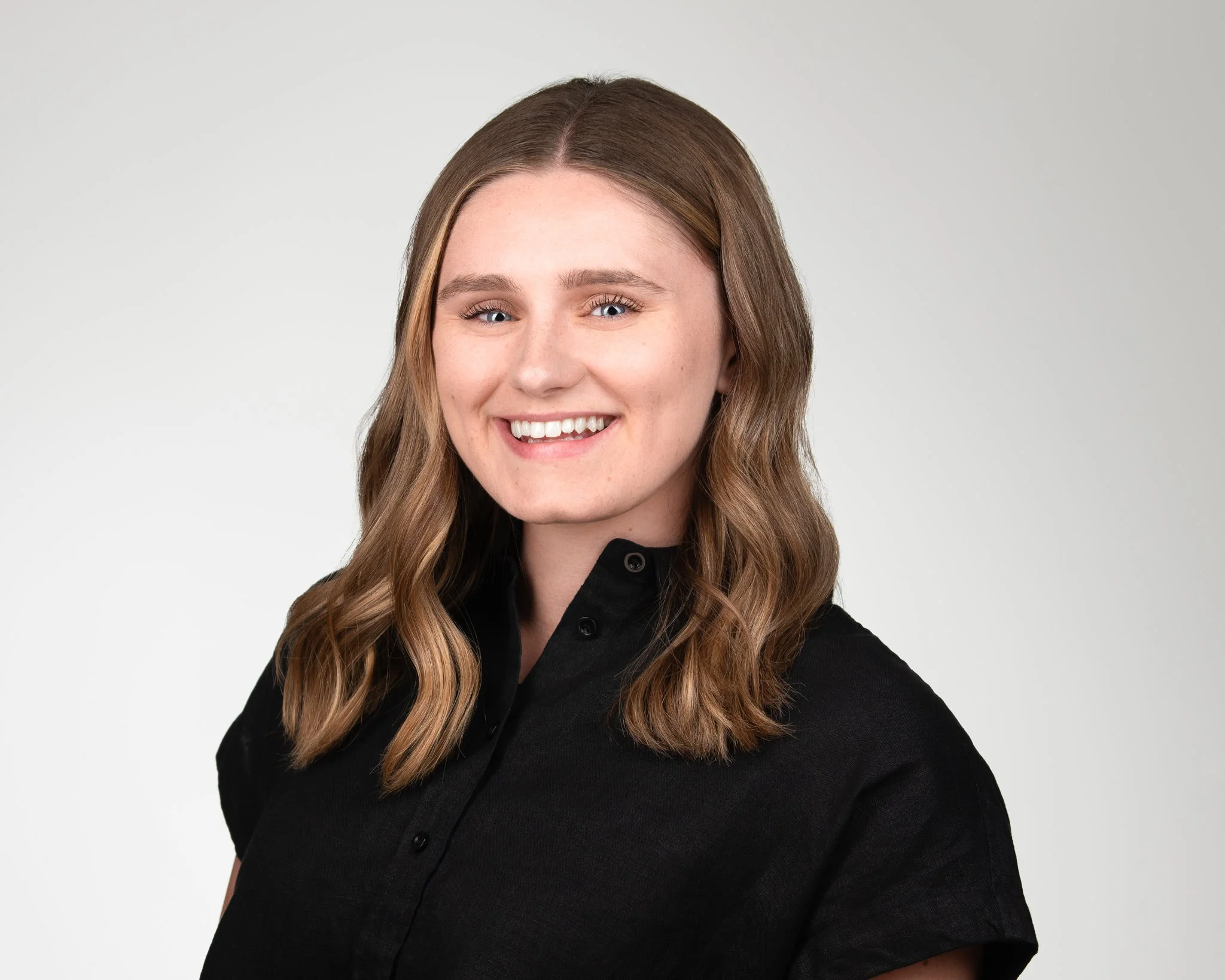 A young woman with light skin and shoulder-length wavy brown hair, smiling and wearing a black shirt, against a plain light gray background.