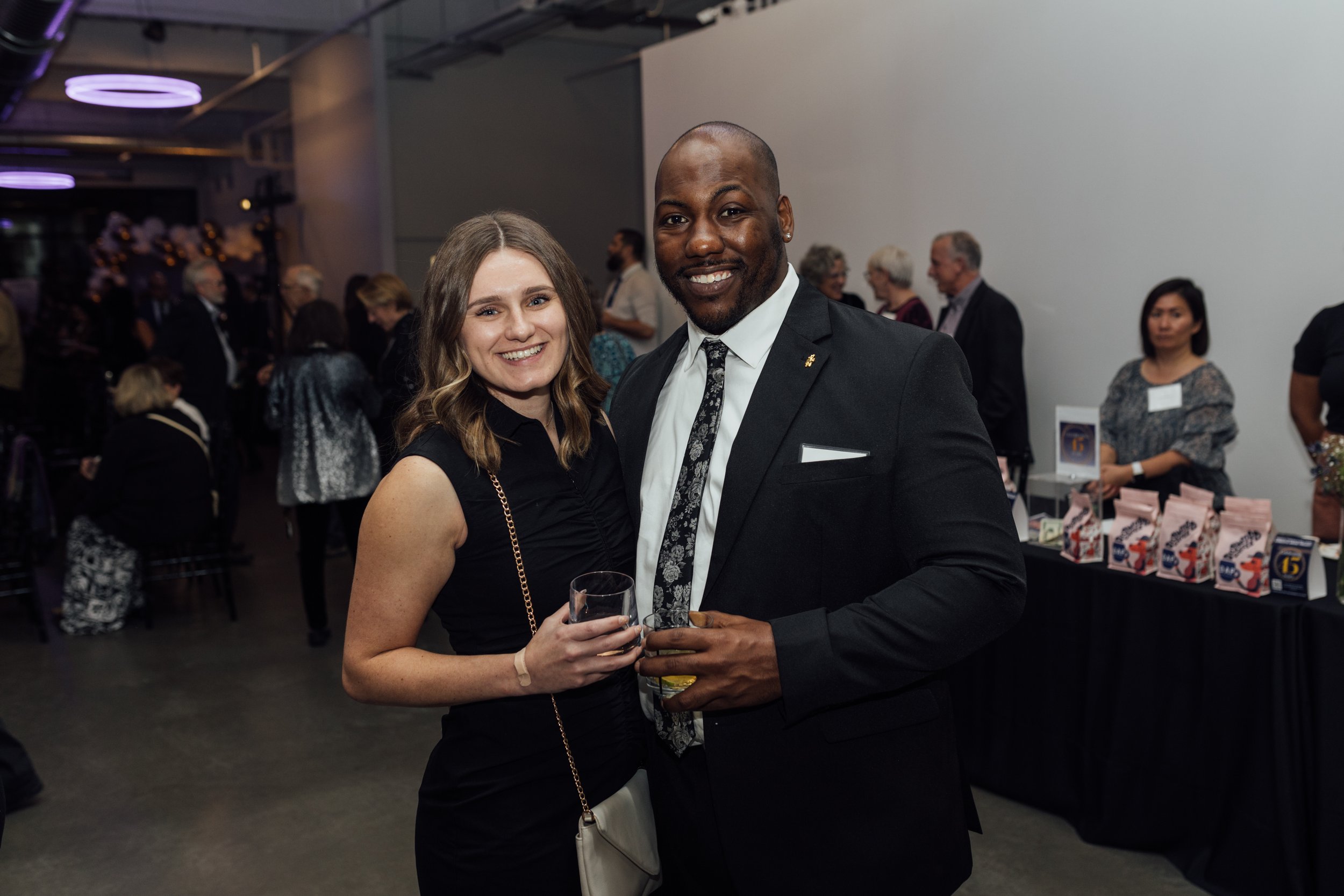 A smiling woman in a black dress stands next to a man in a suit, holding drinks at a social event with people mingling in the background.