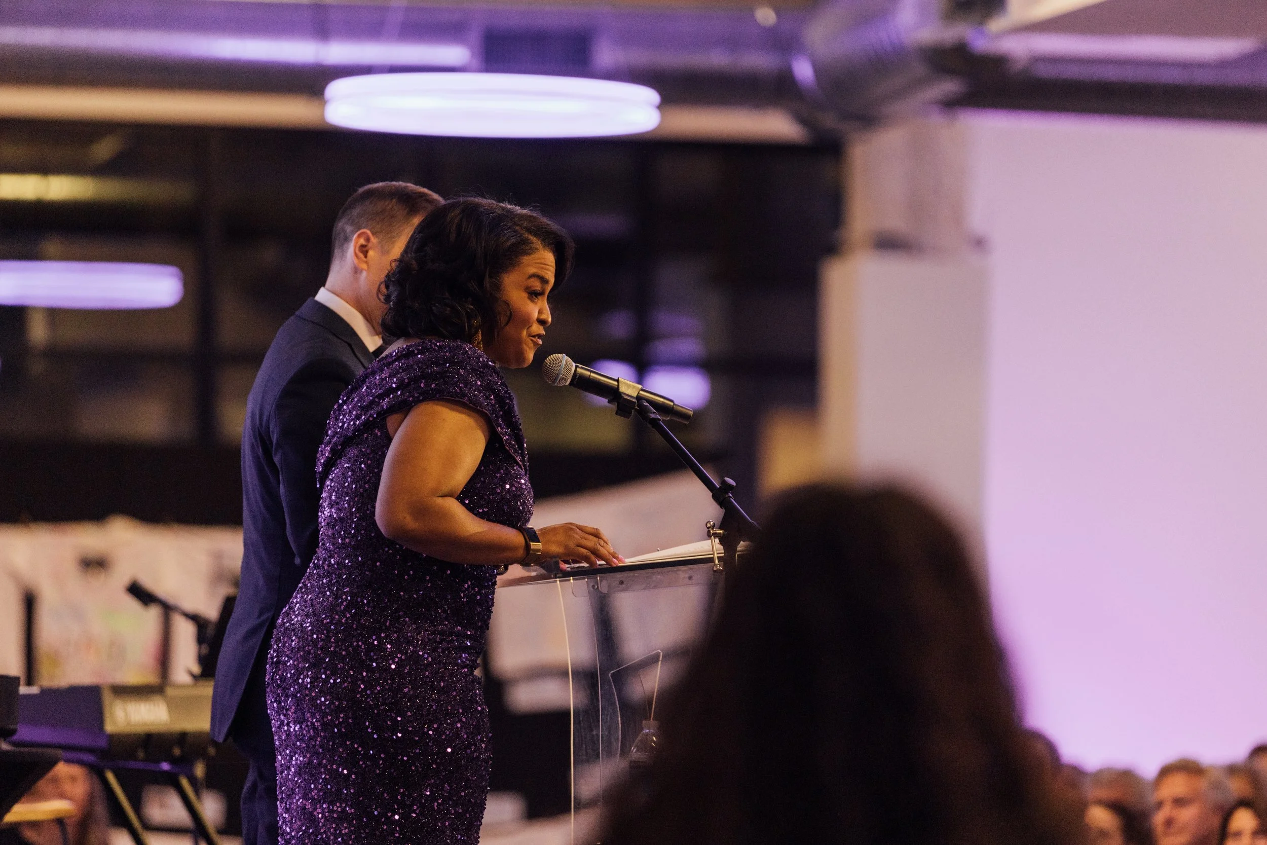 Woman in a purple sparkly dress speaking at a podium, with a man in a dark suit standing behind her, in a room with purple lighting and an audience.