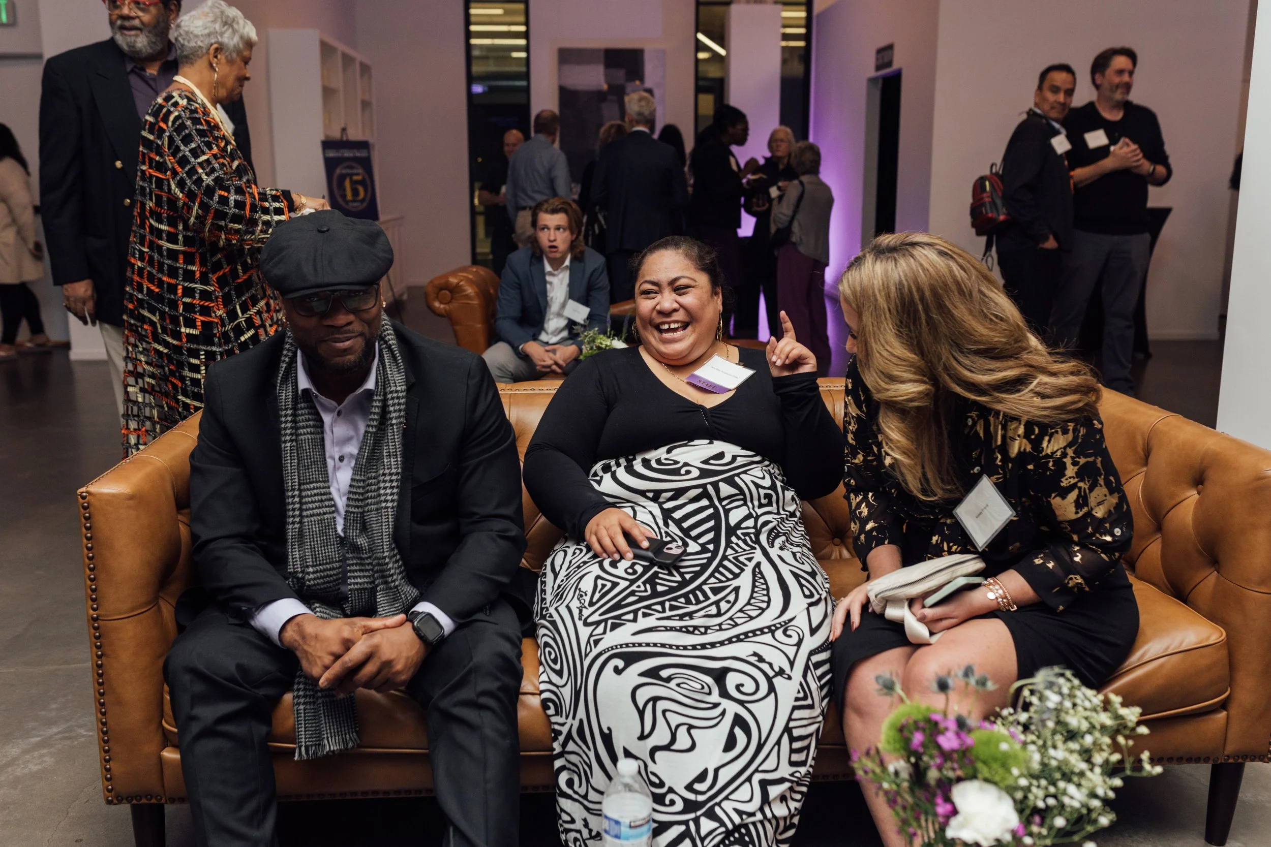Three people sitting on a tan couch at a social event, with additional groups mingling in the background. The person in the middle is smiling and pointing upwards, wearing a black top and a black-and-white patterned skirt, with a cell phone in her ha