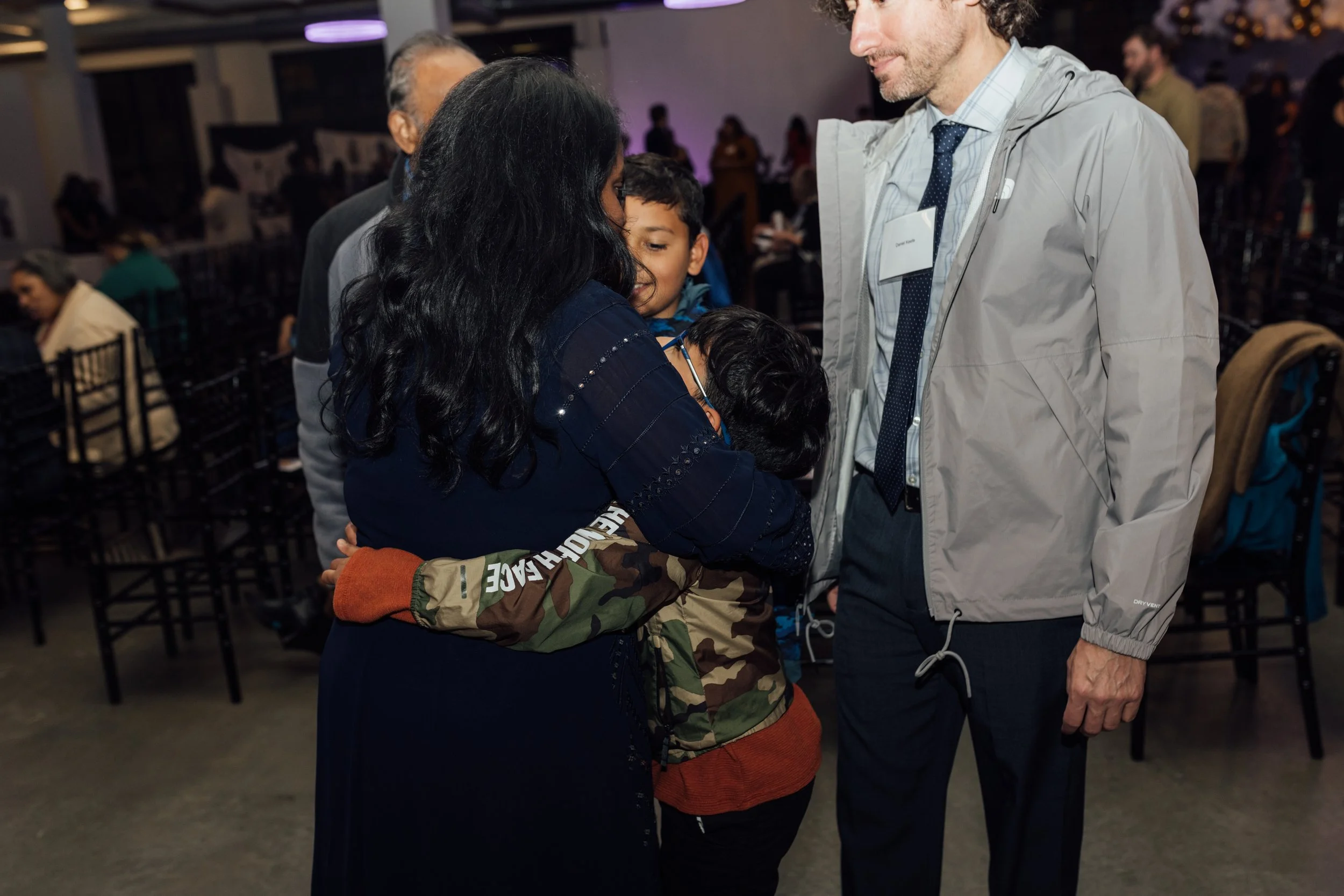 A woman hugging two kids, one in a camouflage jacket, in a crowded indoor event. A man in a gray jacket and tie stands nearby. Others are visible in the background.
