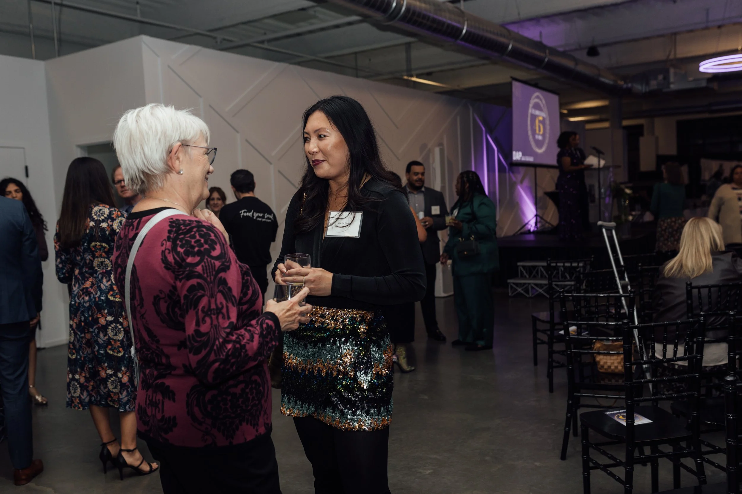 Two women conversing at a formal event, with several other people in the background, some standing and talking, some sitting, in a modern venue with a stage and a large screen displaying a logo.