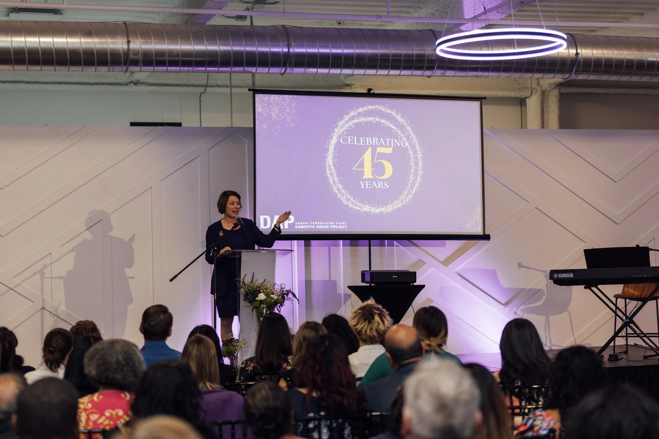 Woman speaking at podium in front of a large screen displaying a 45th-anniversary celebration for the Domestic Abuse Project, with an audience seated watching.