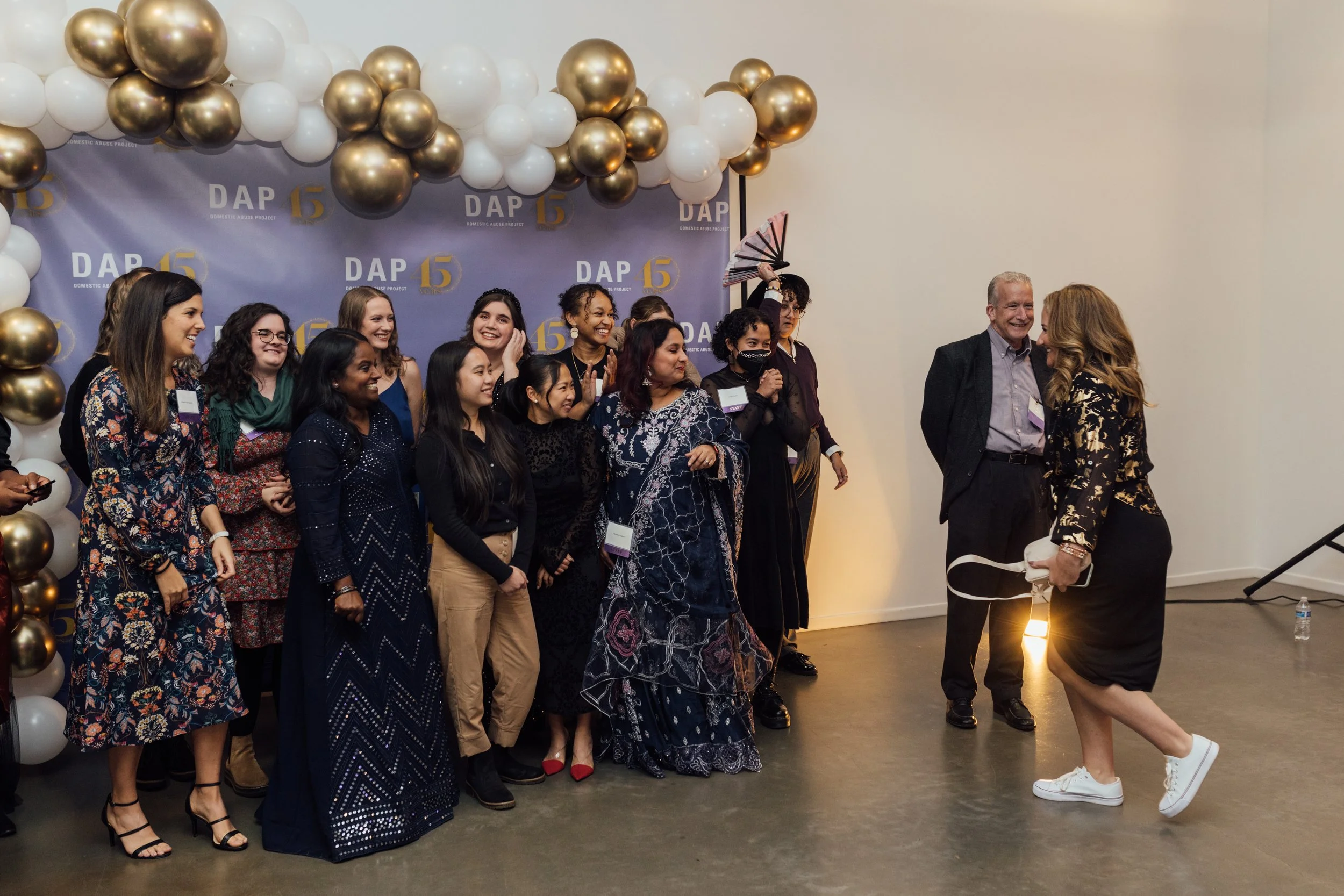 Group of diverse women and two men gathered in front of a backdrop with balloons, celebrating at an event, with some women smiling, chatting, and holding items.